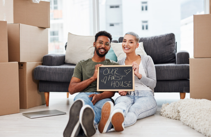 A man and a woman are sitting on the floor holding a sign that says `` our first house ''.