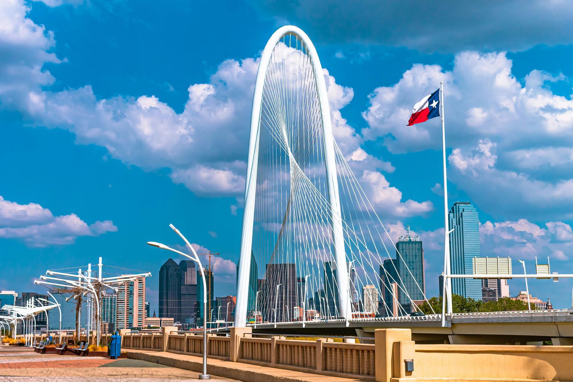 The Margaret Hunt Hill Bridge with its white arch against a blue, cloudy sky and the Dallas city skyline in the distance.