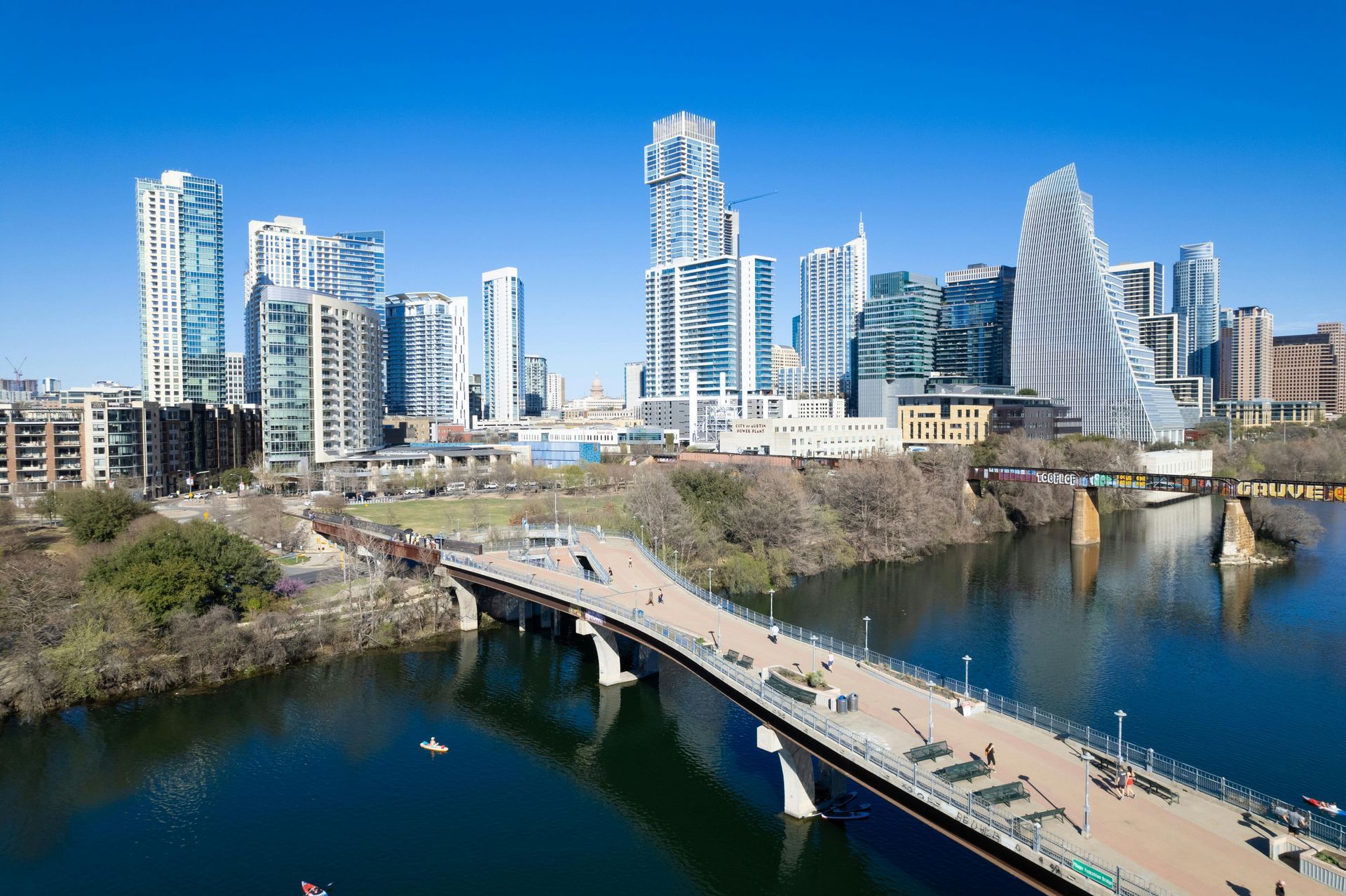 A modern downtown skyline with reflective skyscrapers rising behind a pedestrian bridge crossing a river on a sunny day.