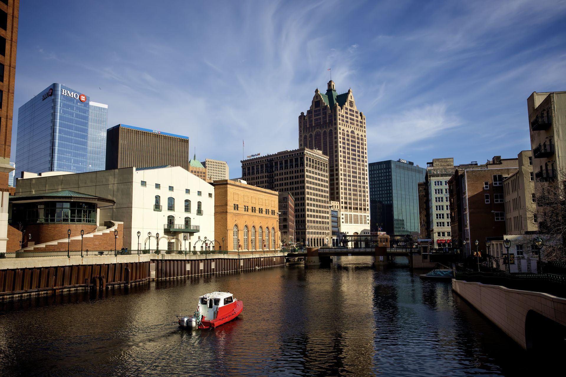 Riverfront city skyline with tall buildings and a small red boat on the water under a blue sky