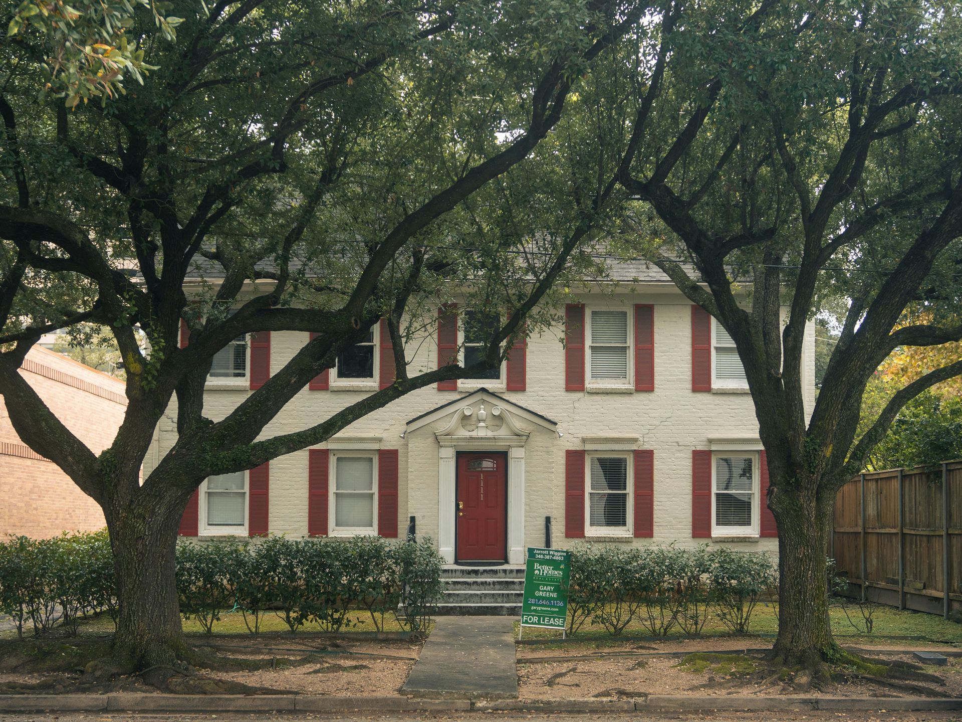 Two-story white brick house with a red door, red shutters, and large trees in the front yard.