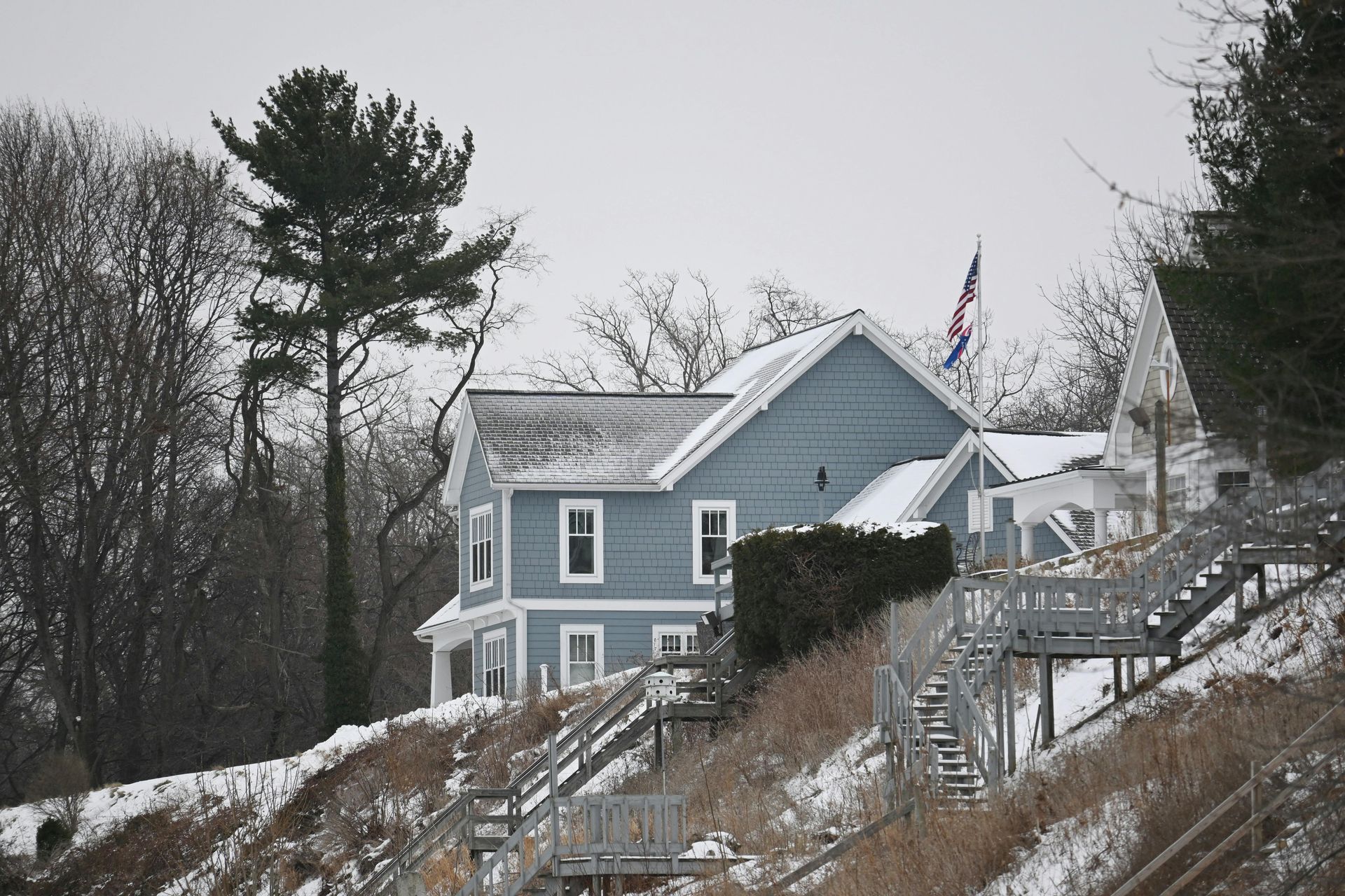 Blue house on a snowy hillside with bare trees and an American flag at dusk