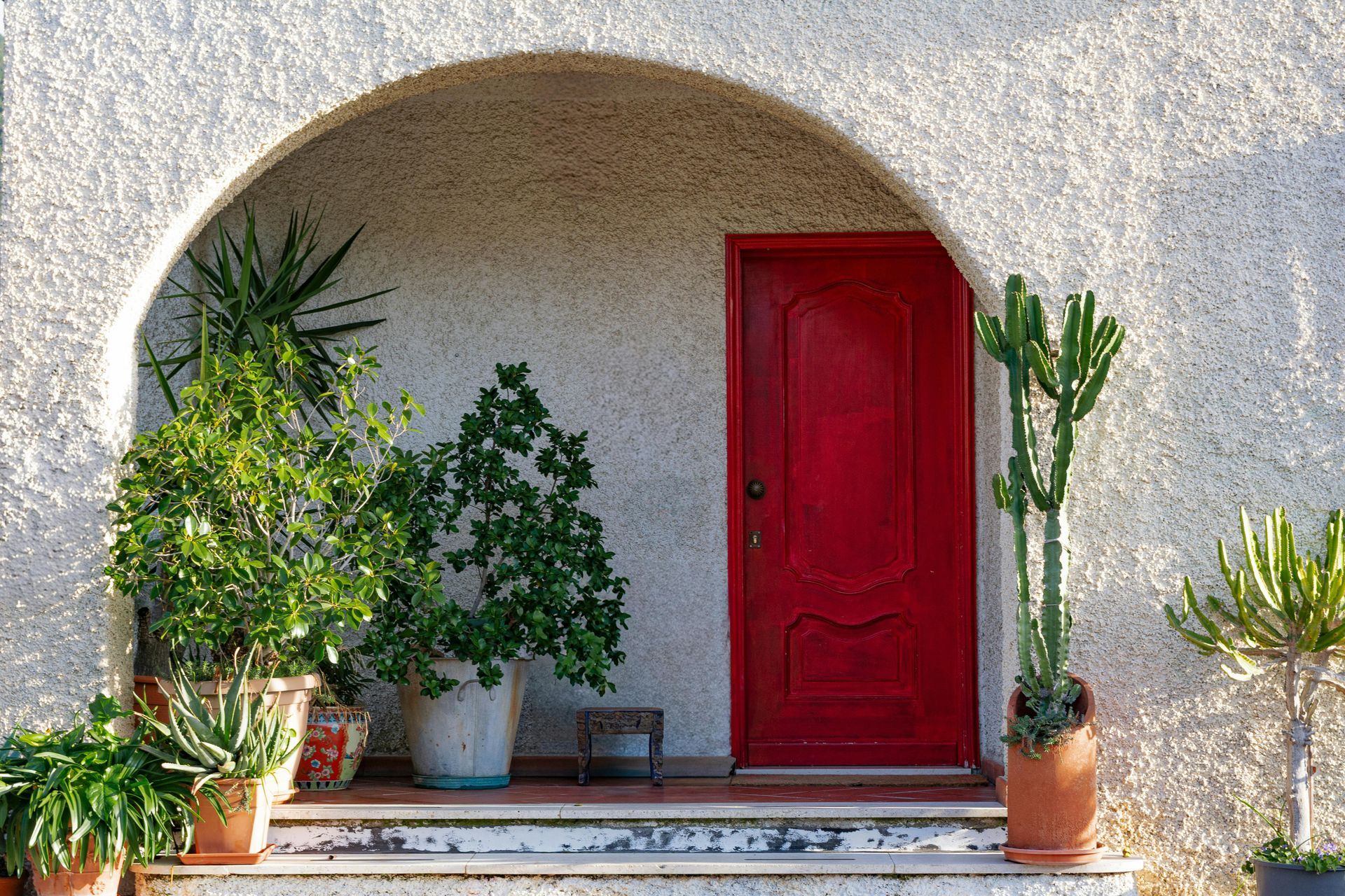 A bright red door is set in a stucco archway, flanked by various potted plants on tiered stone steps.