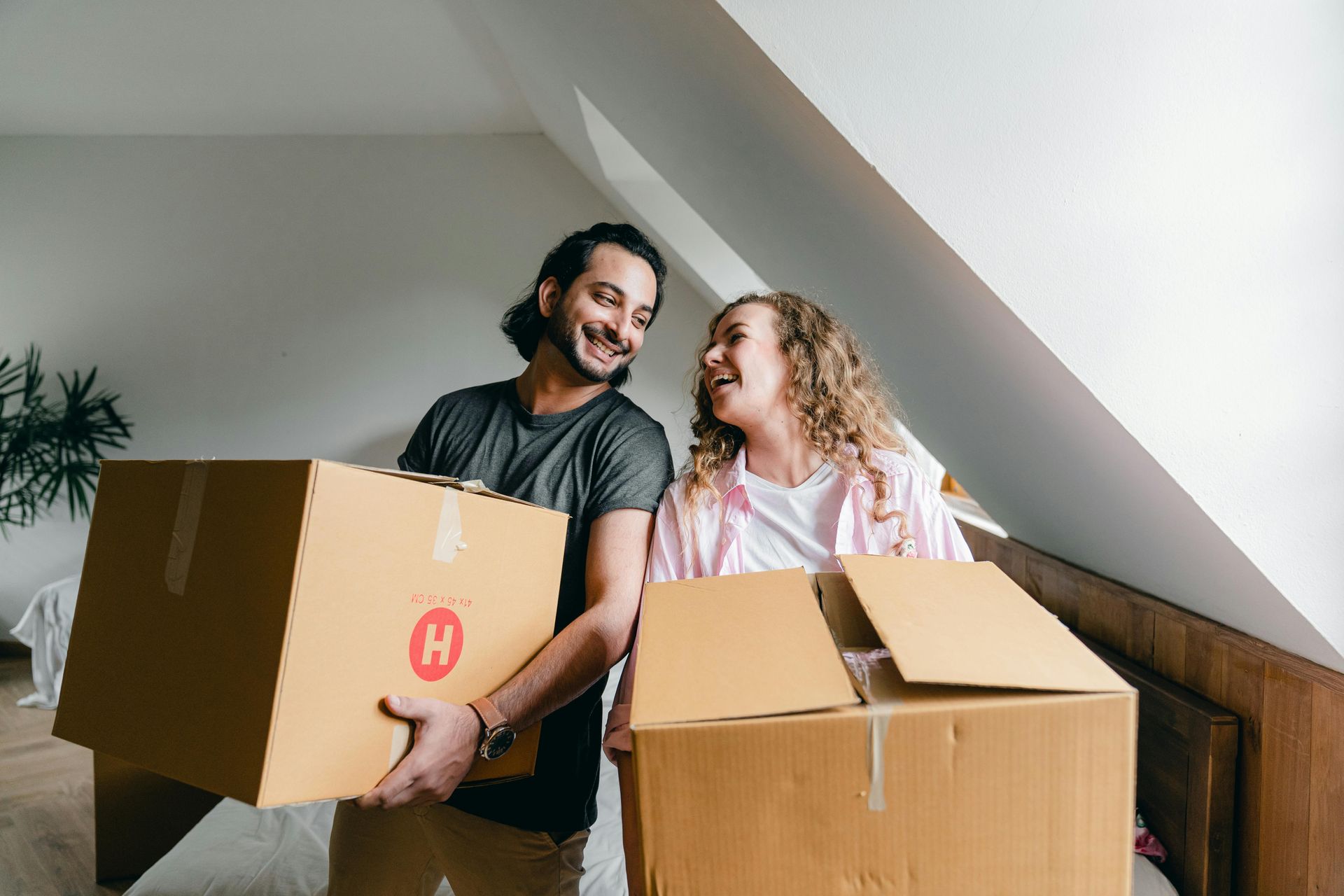 A man and a woman are holding cardboard boxes in a bedroom.