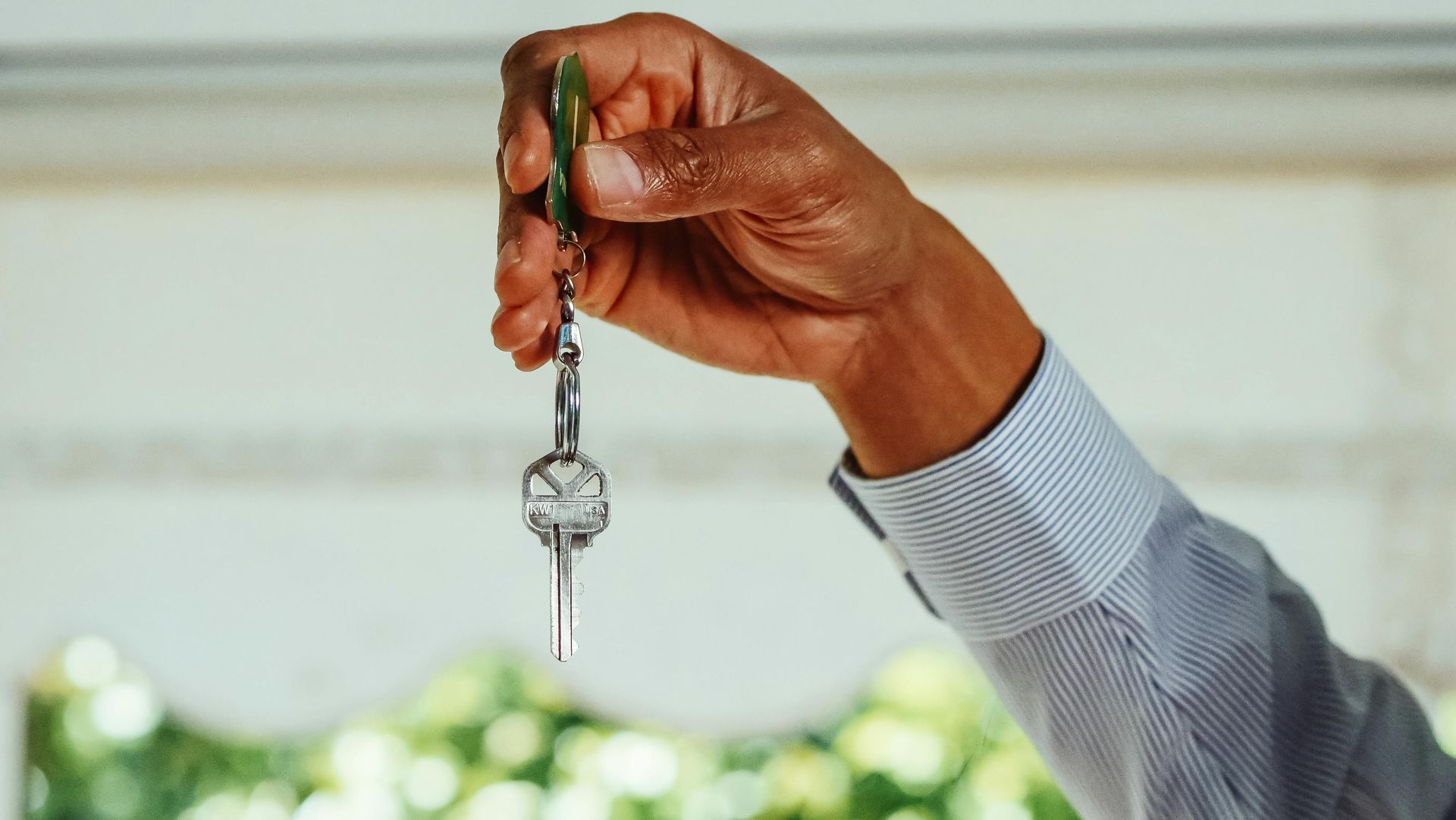 Hand holding keys with a key chain, blurred background of window and greenery.