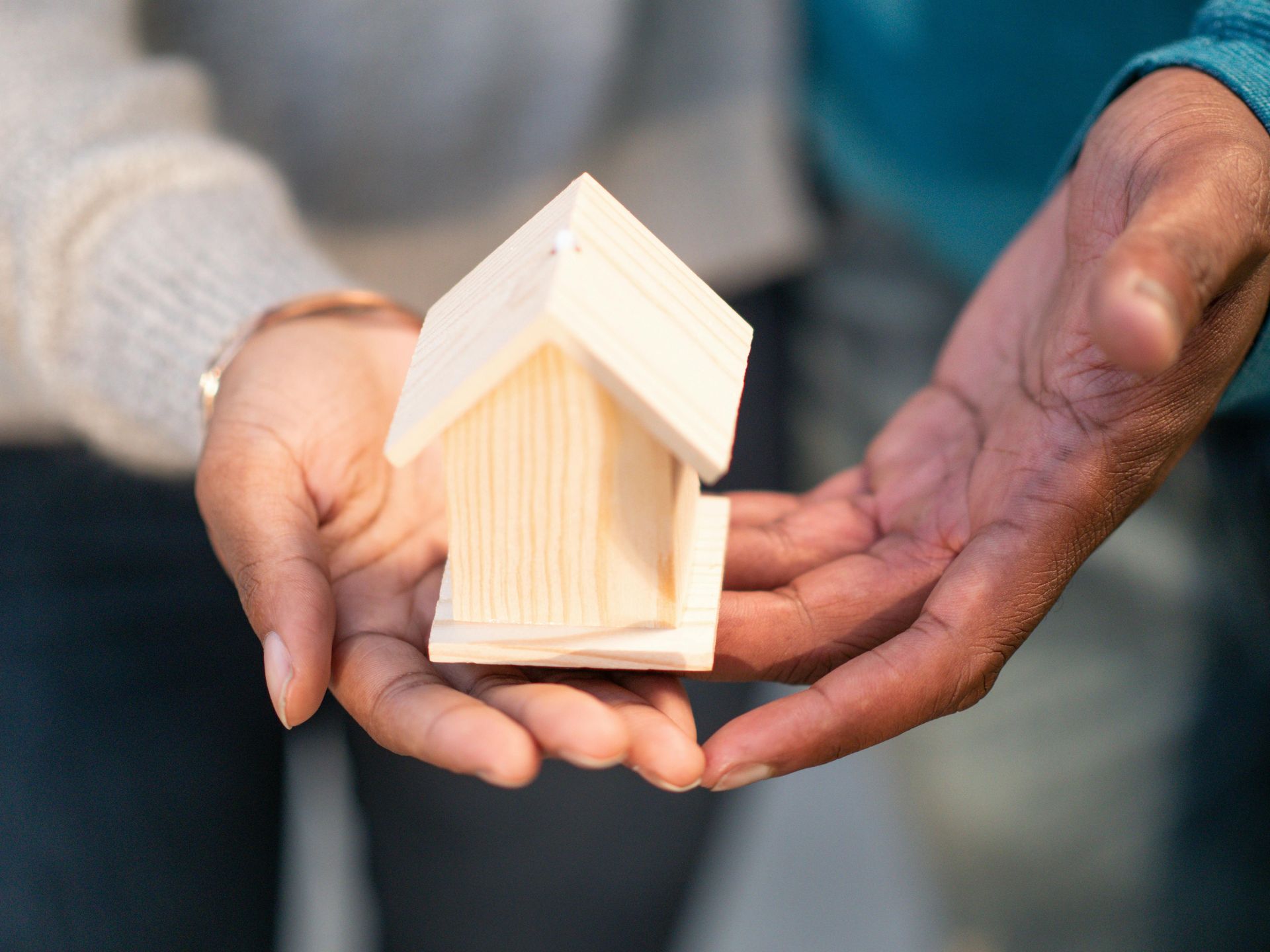 Two sets of hands hold a small, light-colored wooden model of a house together.