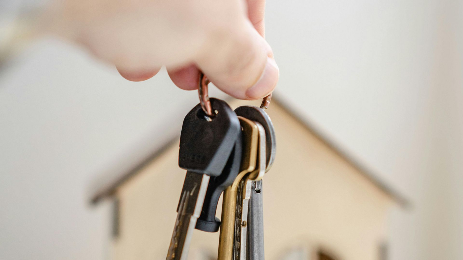 Hand holding a key ring with keys in front of a miniature wooden house.