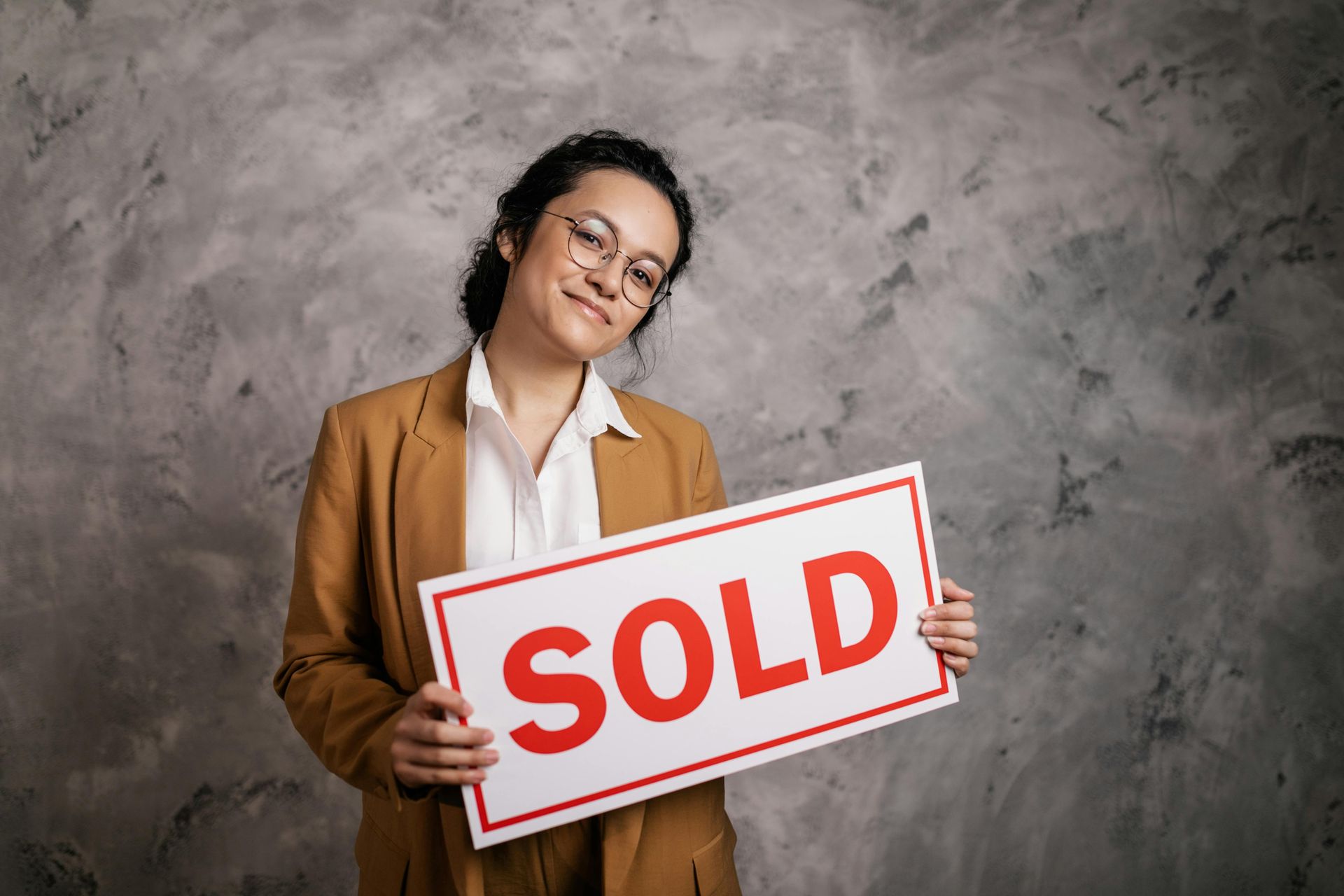 Person holding a red and white “SOLD” sign against a gray backdrop, smiling with eyes closed