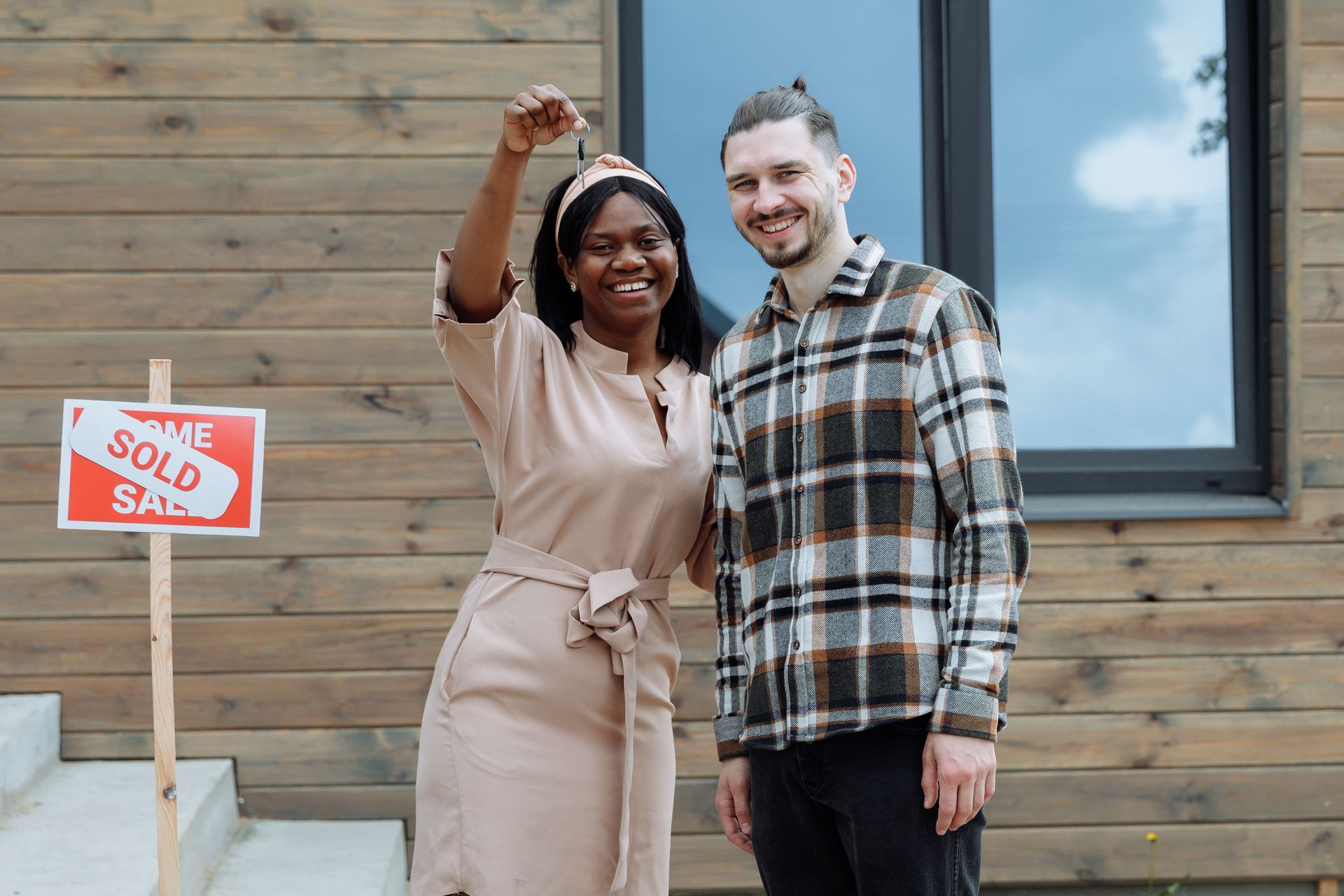 Two people smiling and holding keys in front of a house with a sold sign