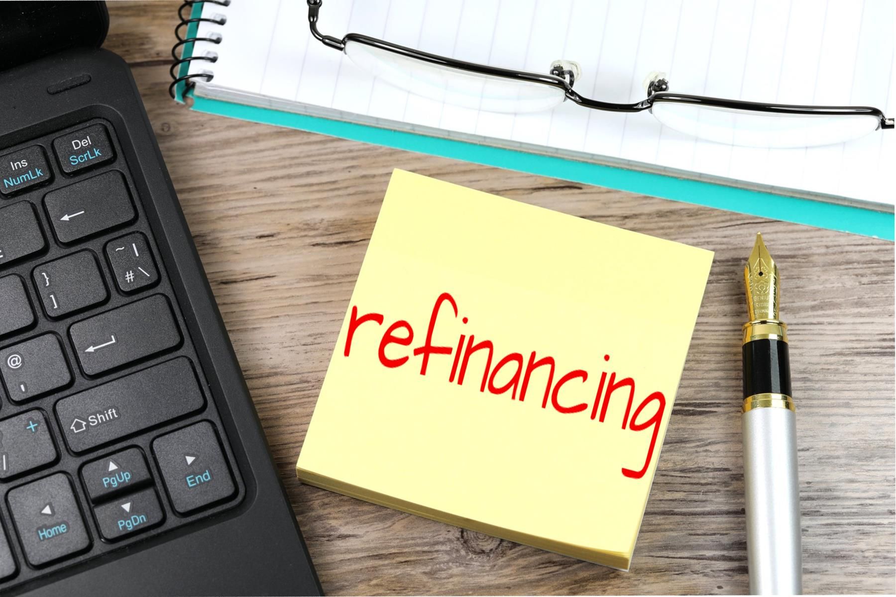 A sticky note with the word refinancing written on it is next to a laptop and a pen.