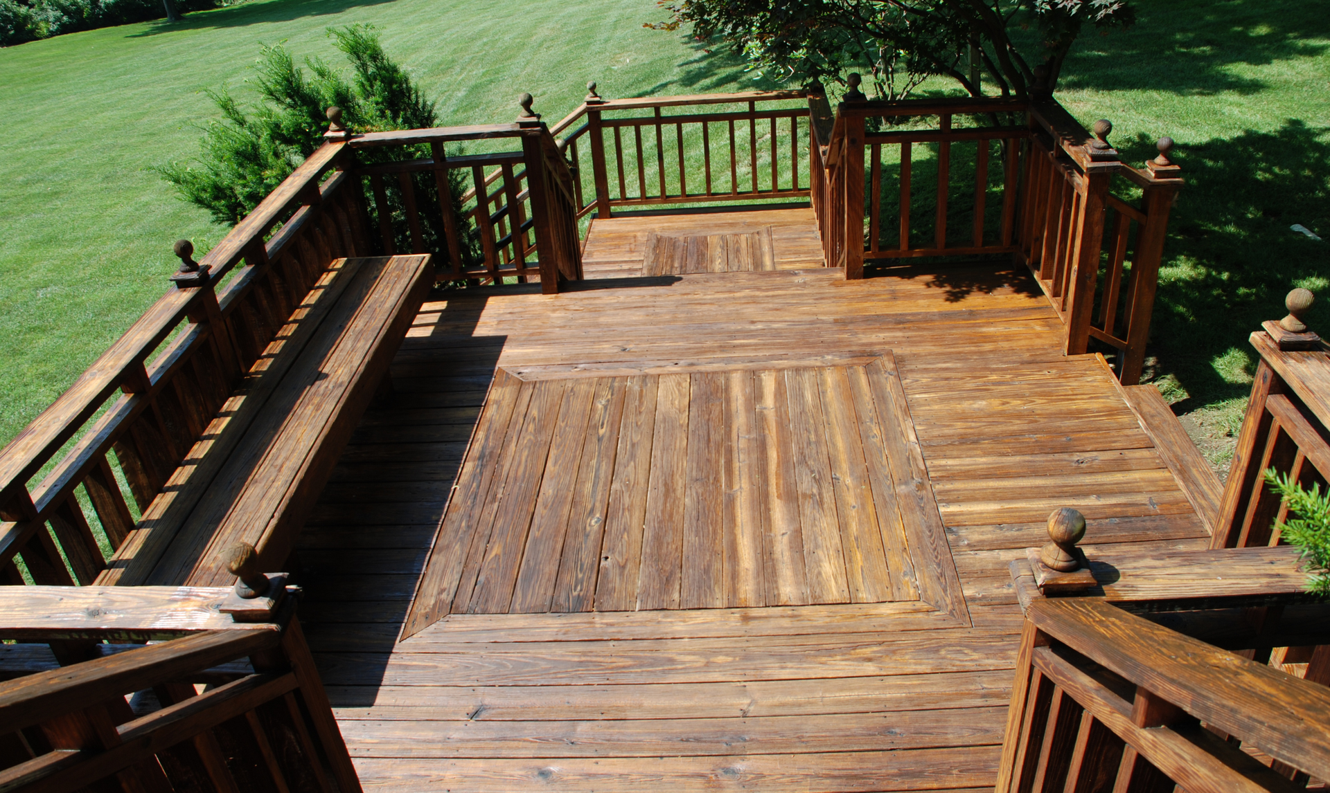 Wooden deck with railings and bench, surrounded by green grass and trees.