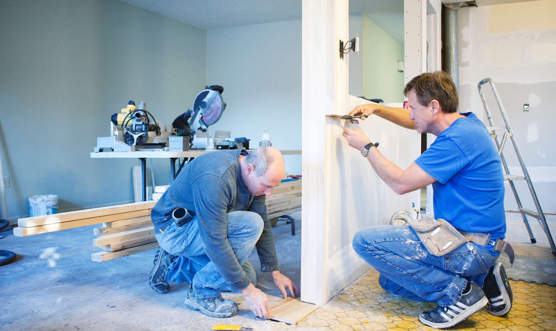 Two men renovating a room, one installing trim, the other working on flooring.