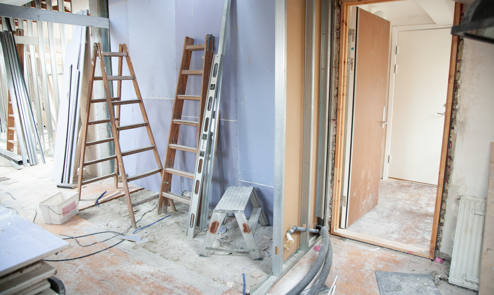 Room under renovation with ladders and tools; doorframe open to another room.