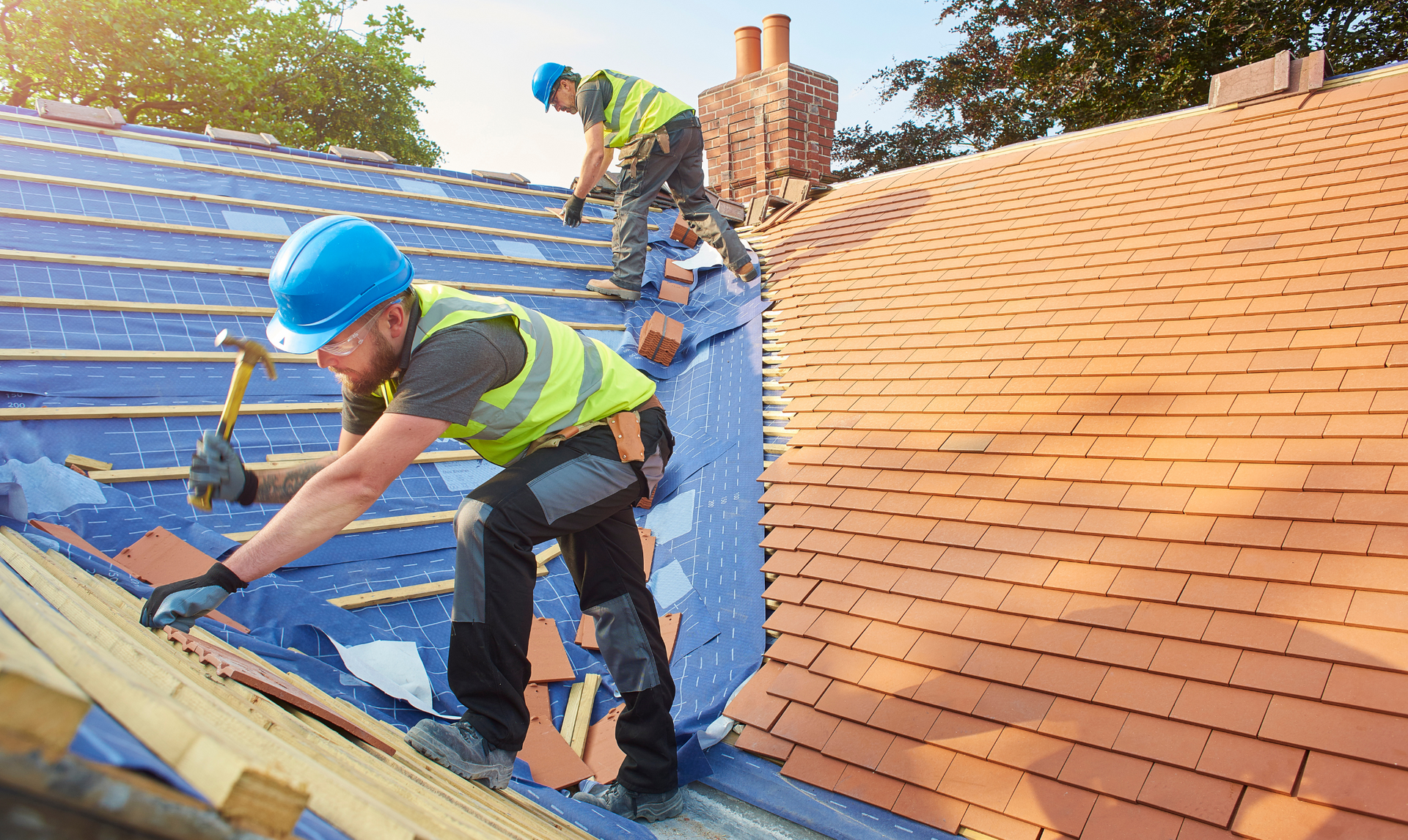 Two roofers in hard hats and vests working on a tiled roof, one hammering, the other nearby.