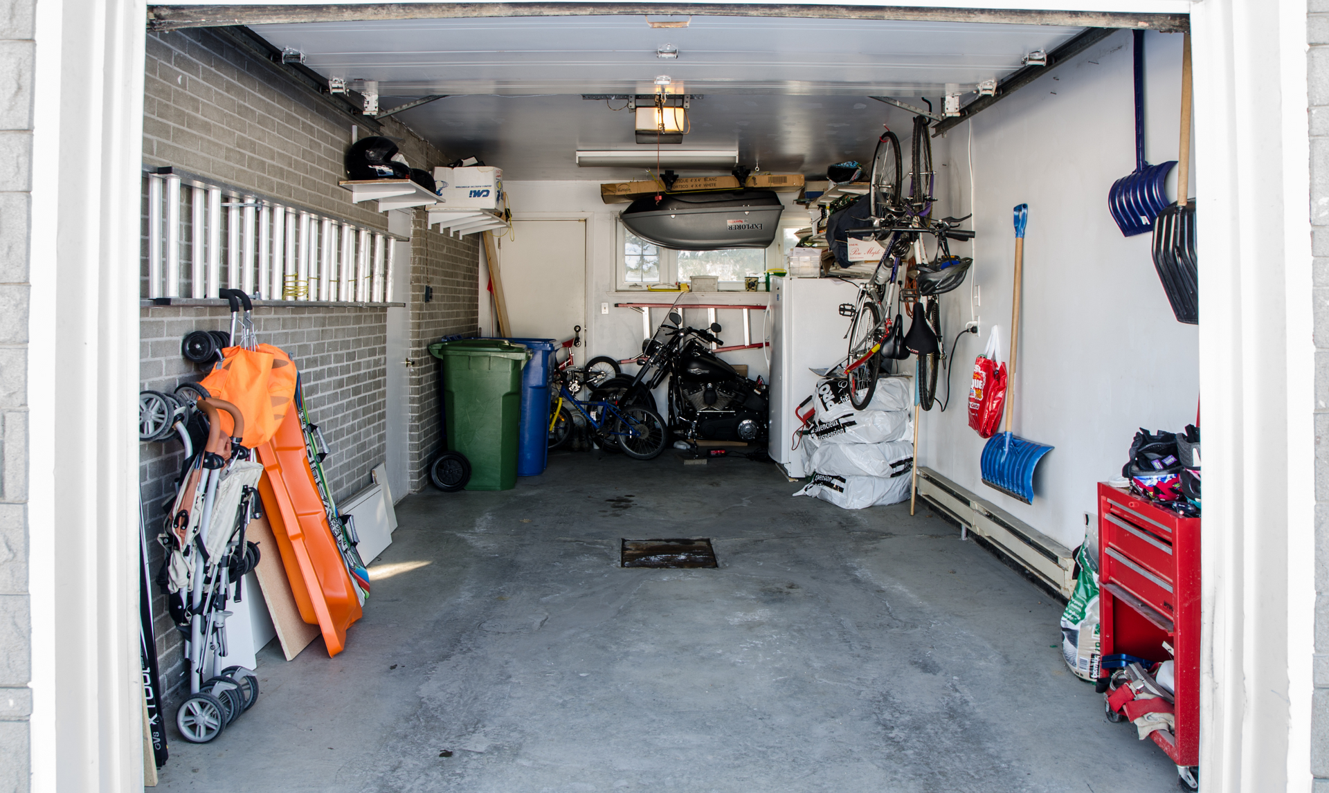 Cluttered garage with bicycles, tools, and storage. Gray concrete floor, white walls, and open doorway.