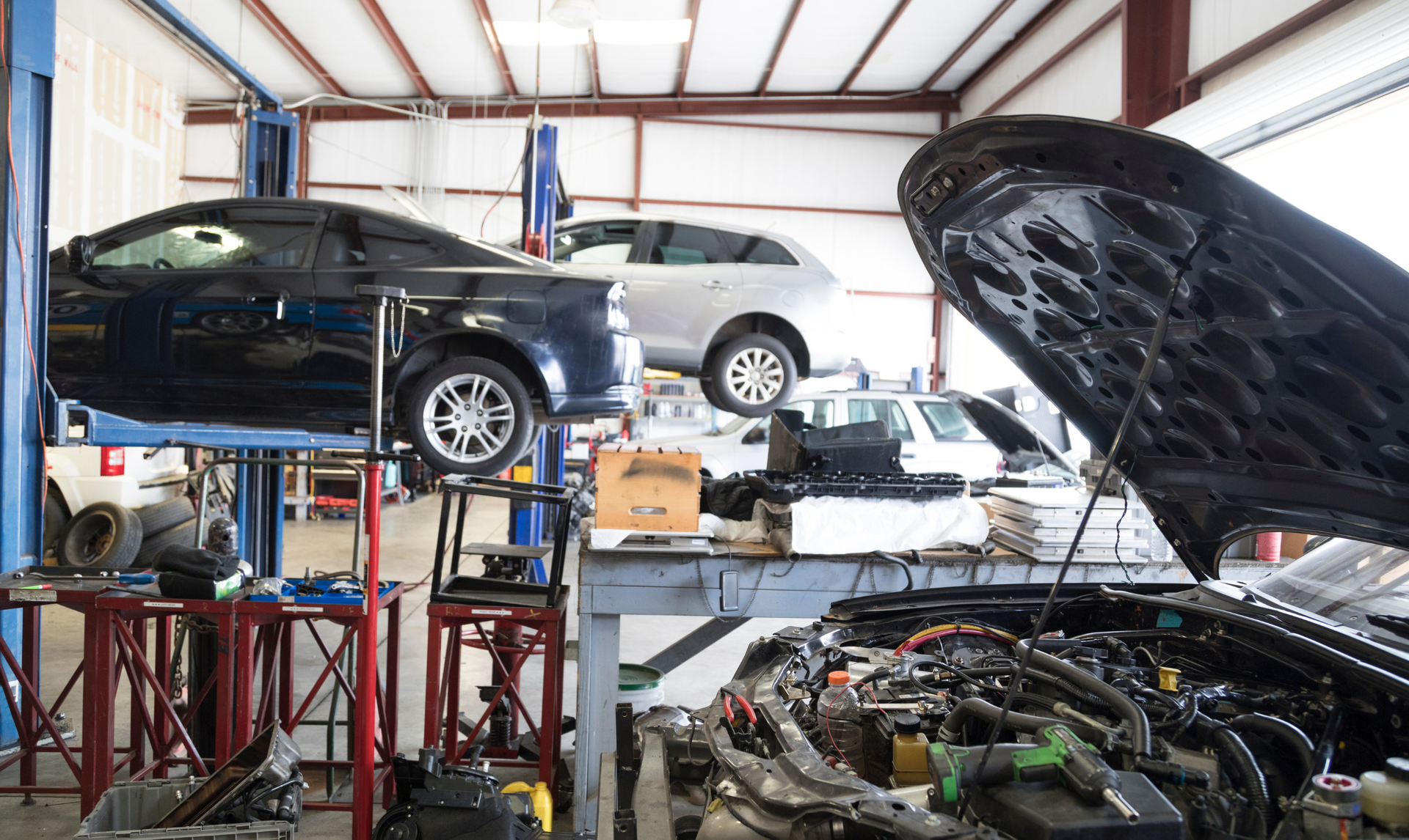 Cars on lifts in a garage; one with hood open. Tools and equipment scattered on work surfaces.