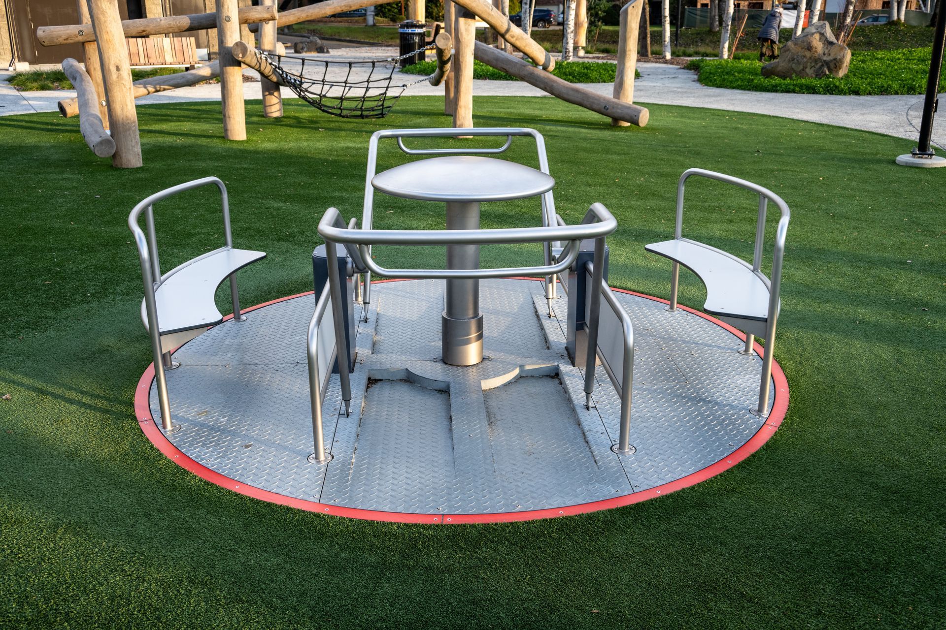 Metal merry-go-round with seats in a playground, set on green turf. Wooden play structures are in the background.