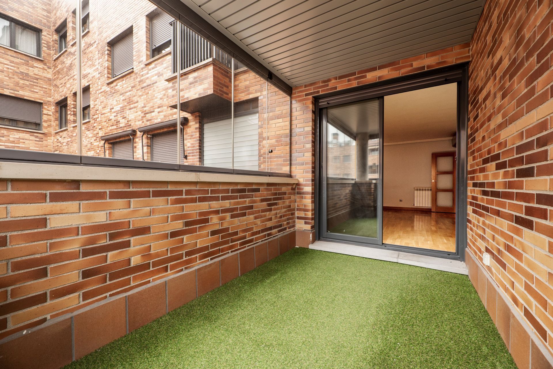 Balcony with brick walls, artificial turf, and sliding glass door leading to an interior with hardwood floors.