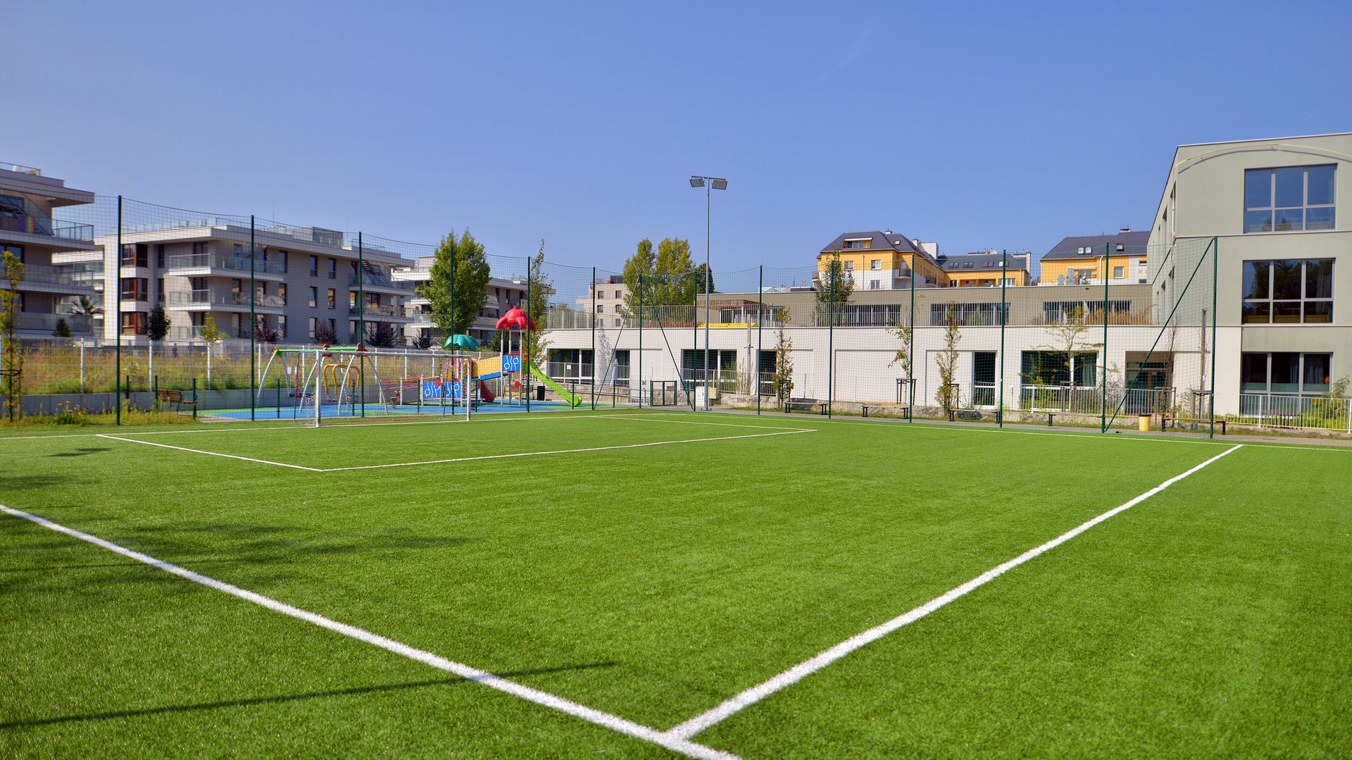 Green artificial turf sports field with white lines, playground, and buildings in the background.
