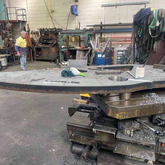 A worker stands in a machine shop beside a large, flat steel plate being prepared on industrial equipment.