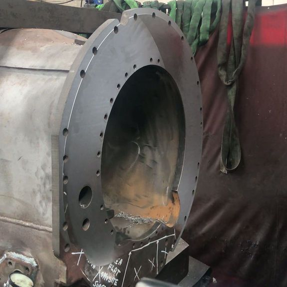 A metal industrial casing with a circular flange featuring pre-drilled bolt holes, resting in a workshop environment.