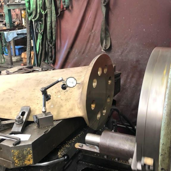 A dial indicator mounted on a metal lathe checks the alignment of a large, circular metal flange with eight holes.
