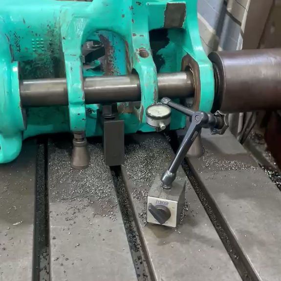 A dial indicator on a magnetic base measures a cylindrical metal shaft set within a green cast iron machinery housing.