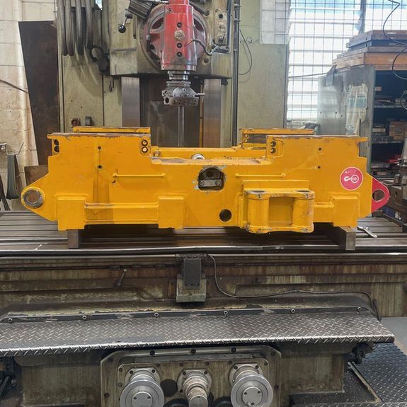 A large yellow industrial steel component sits centered on the metal bed of a milling machine in a workshop.