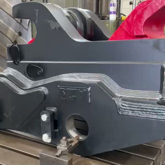 A large, gray industrial steel component with visible welding seams, secured on a machine worktable for fabrication.