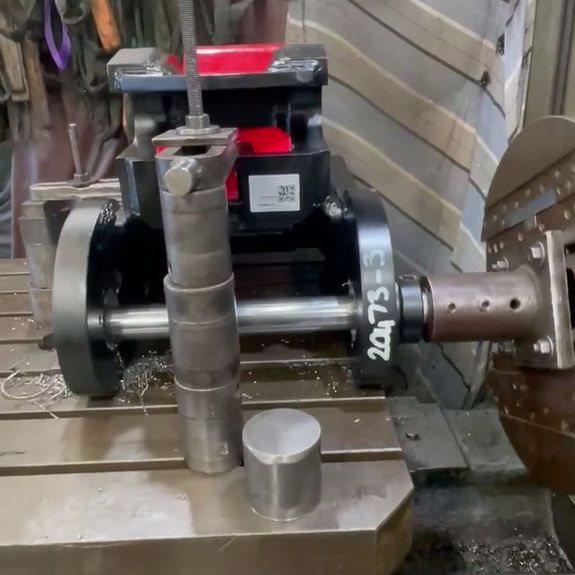 A machined metal part is secured on a milling machine table with a cylindrical spacer sitting in the foreground.