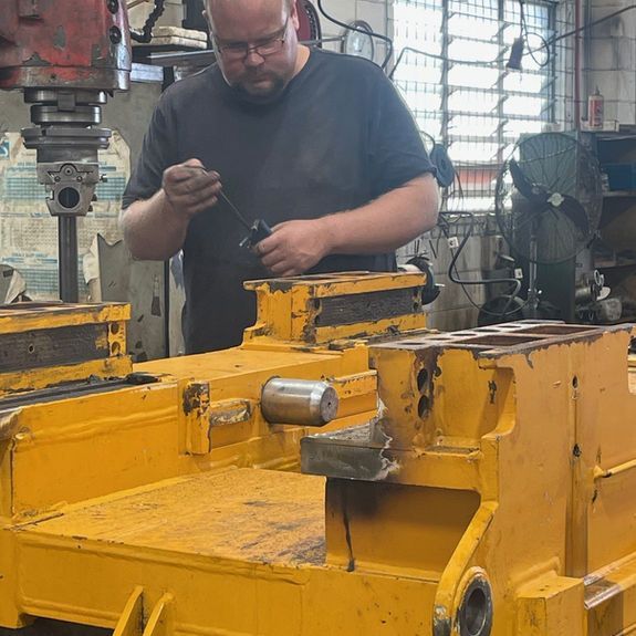 A technician in a workshop inspects a large yellow industrial engine block while standing beneath a heavy-duty drill press.