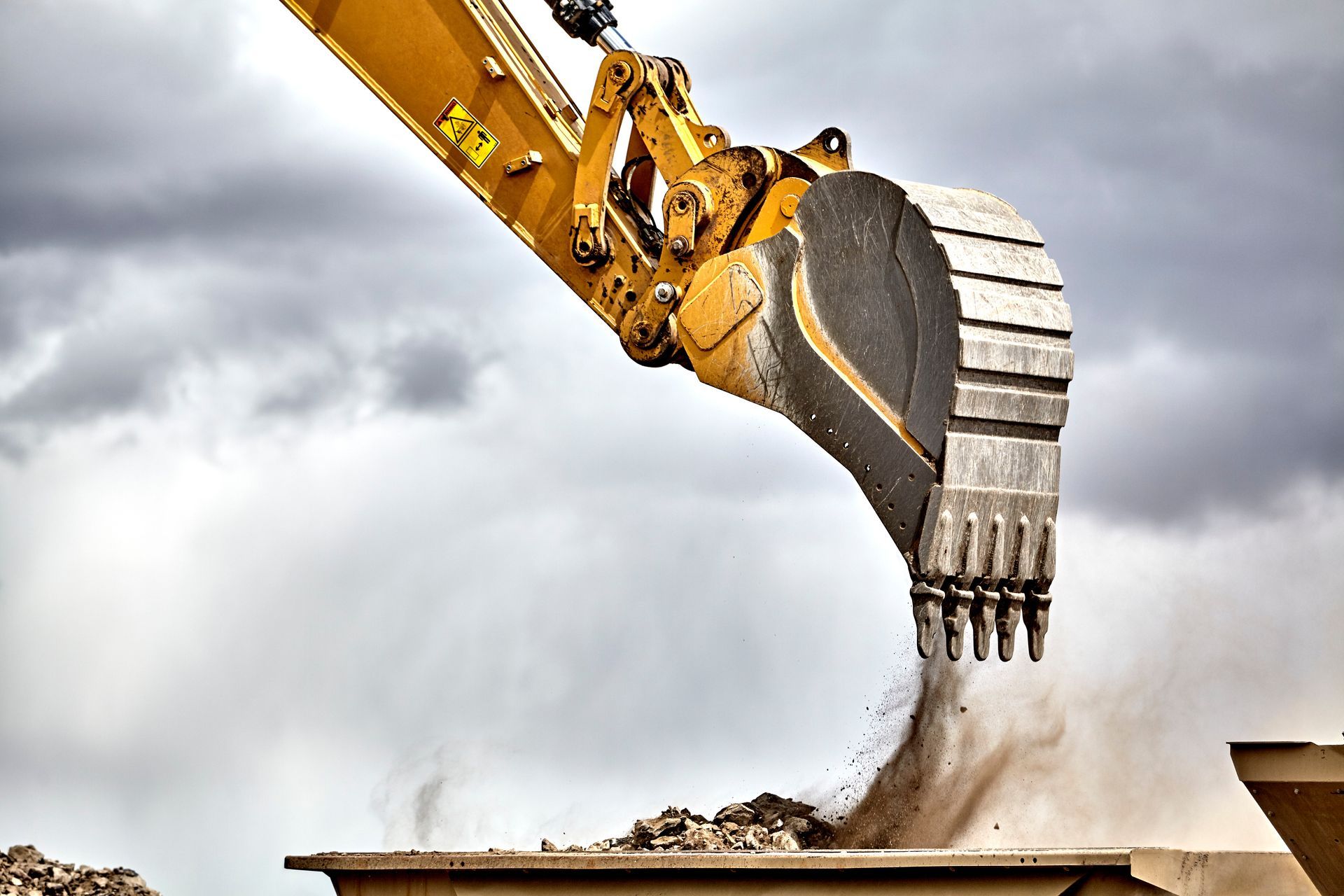 A yellow excavator bucket dumps dirt and rocks into a container against a cloudy sky.