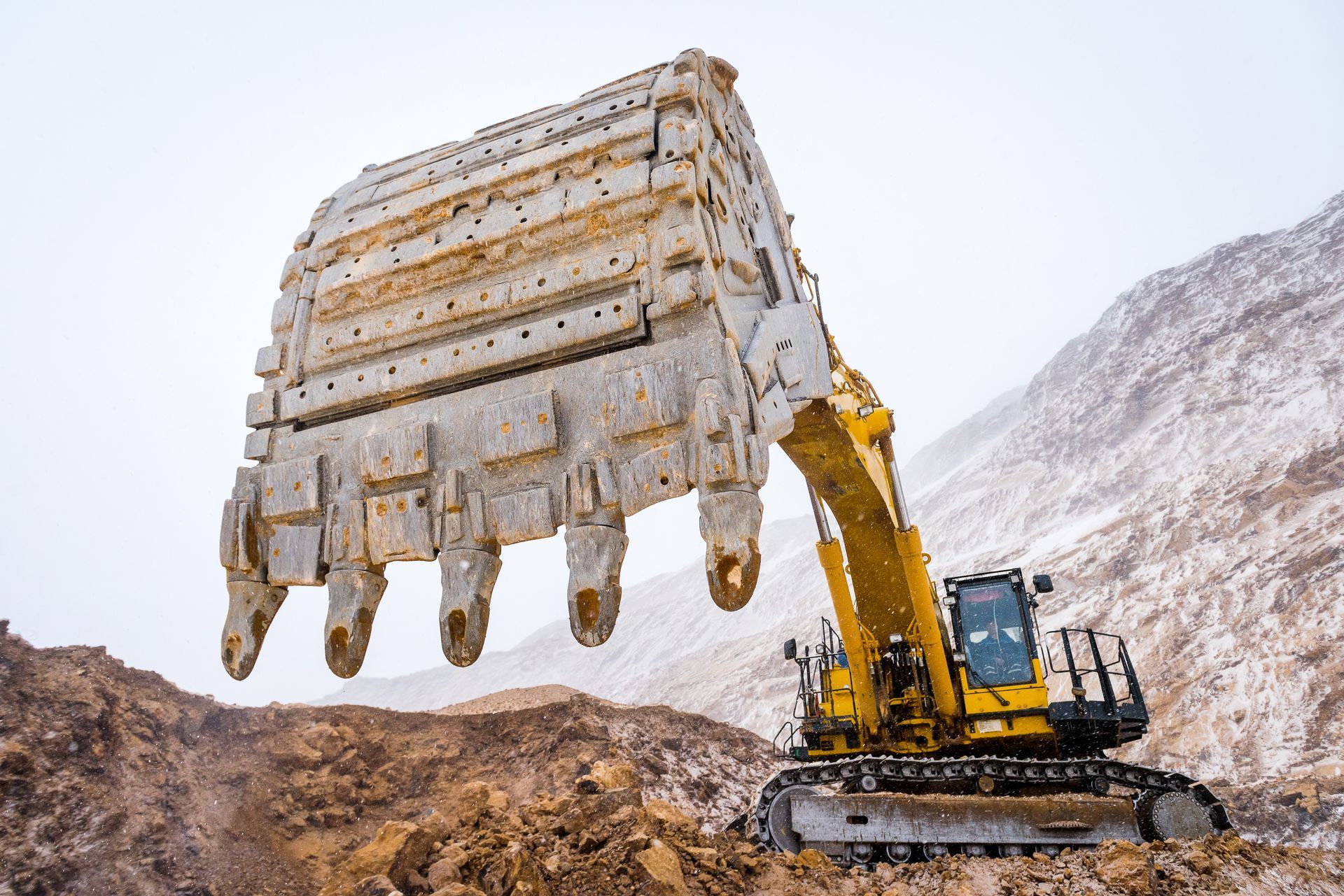 A large yellow excavator raises its heavy-duty bucket against a backdrop of a snowy, rocky mining landscape.