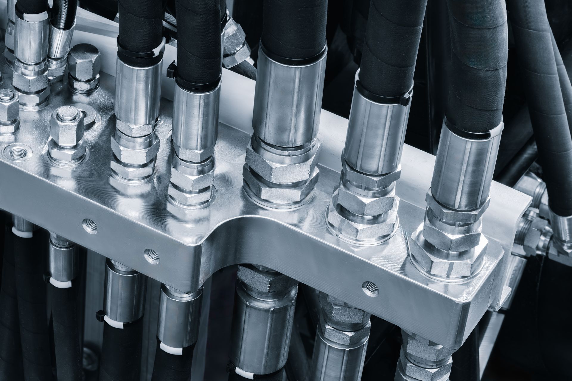 Close-up of a metal industrial manifold block with multiple black hydraulic hoses connected by silver couplings.
