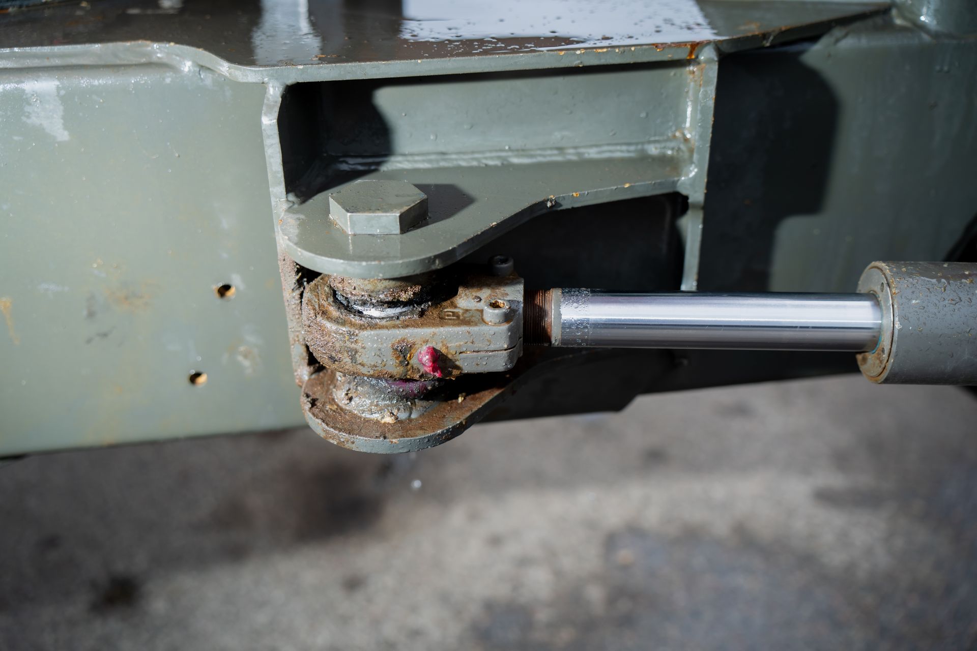 A metal clevis joint connects a hydraulic cylinder to a painted green frame with a bolt, showing signs of wear and rust.