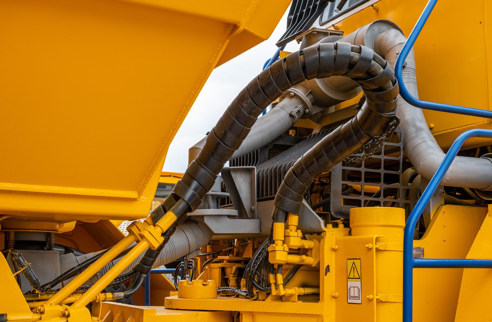 A close-up of heavy construction machinery featuring yellow panels, a protective black hose sleeve, and blue metal railing.