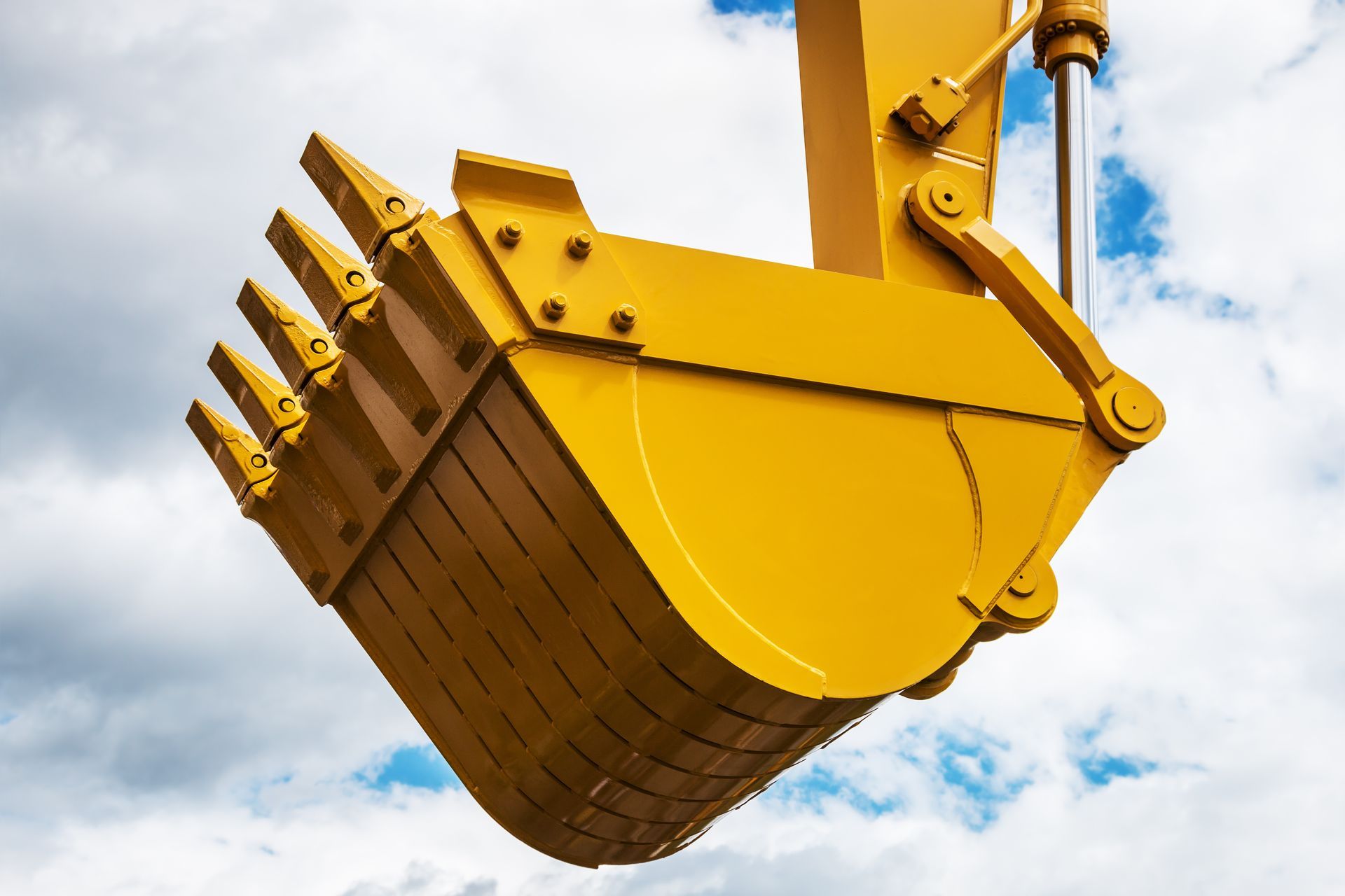 A bright yellow excavator bucket with metal teeth, positioned against a cloudy blue sky.