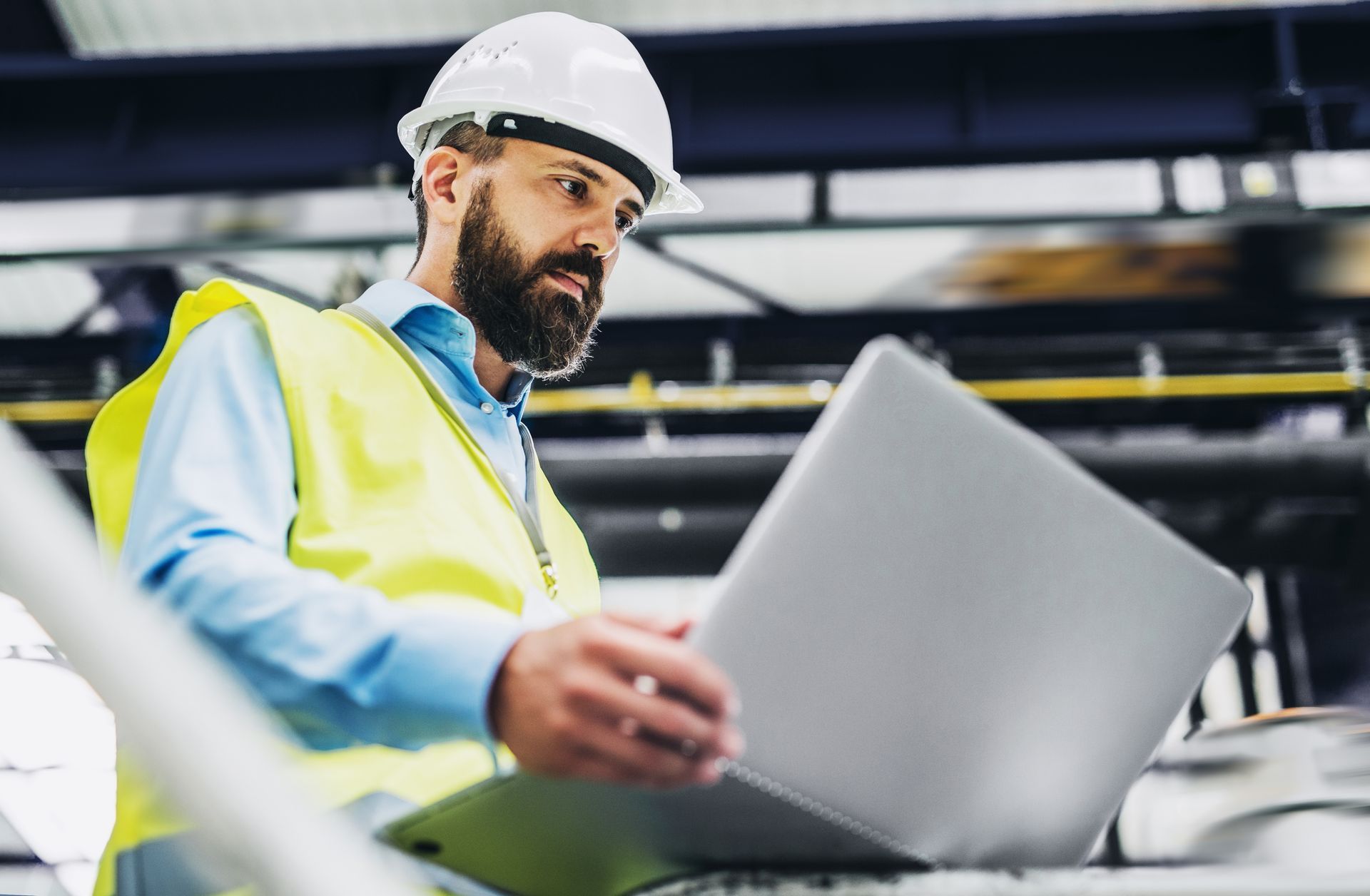A person in a high-visibility vest and hard hat looking at a laptop in an industrial setting.