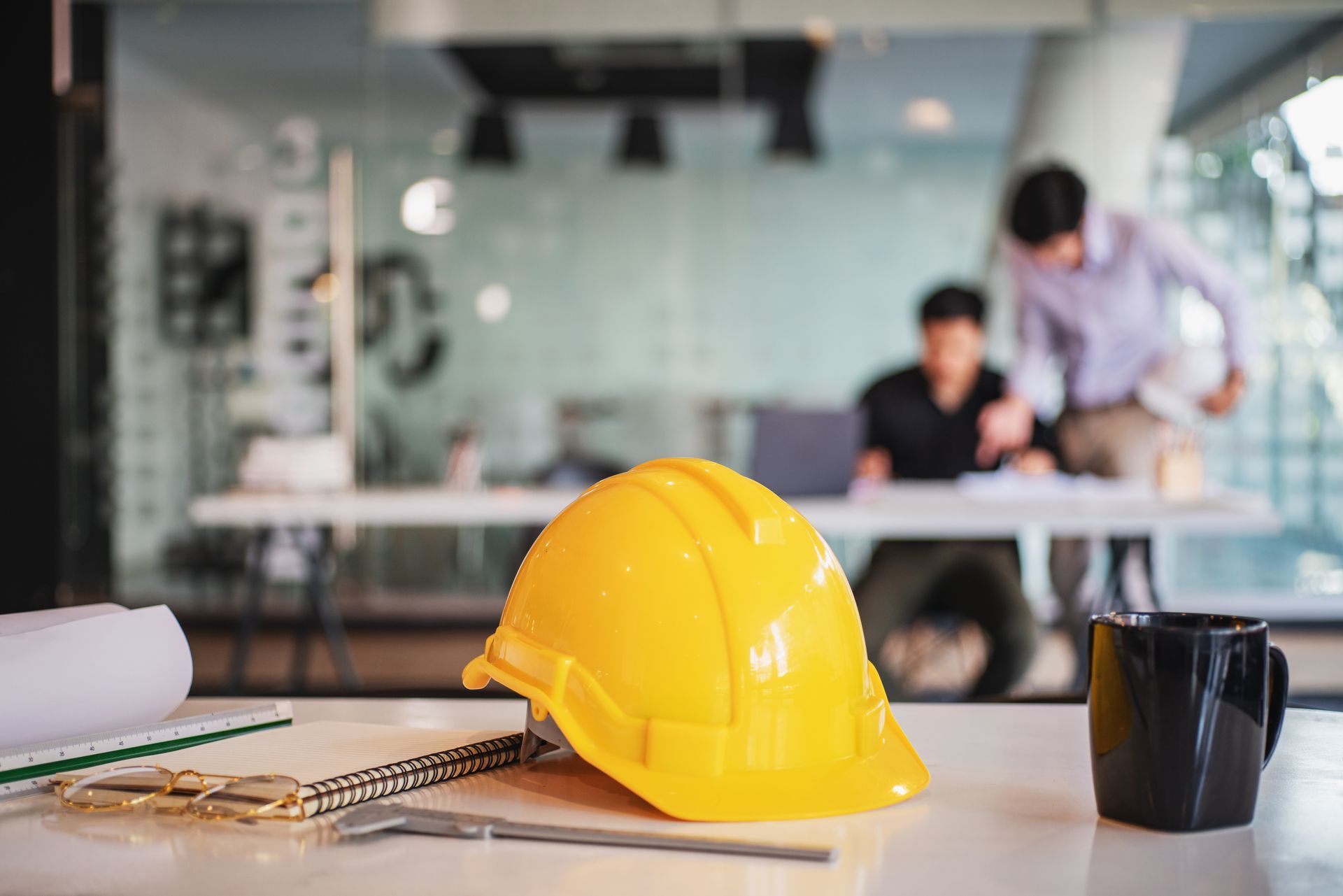 A yellow hard hat sits on a desk next to tools and a mug, with two people working in a blurred office background.