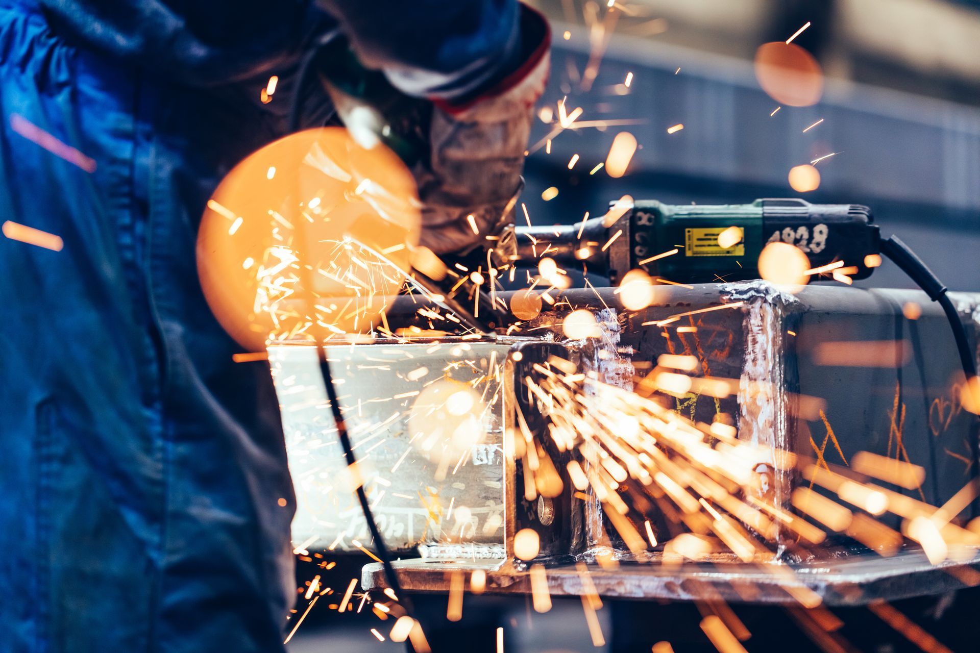 A worker wearing blue trousers uses a power tool to grind a metal object, producing a spray of bright orange sparks.