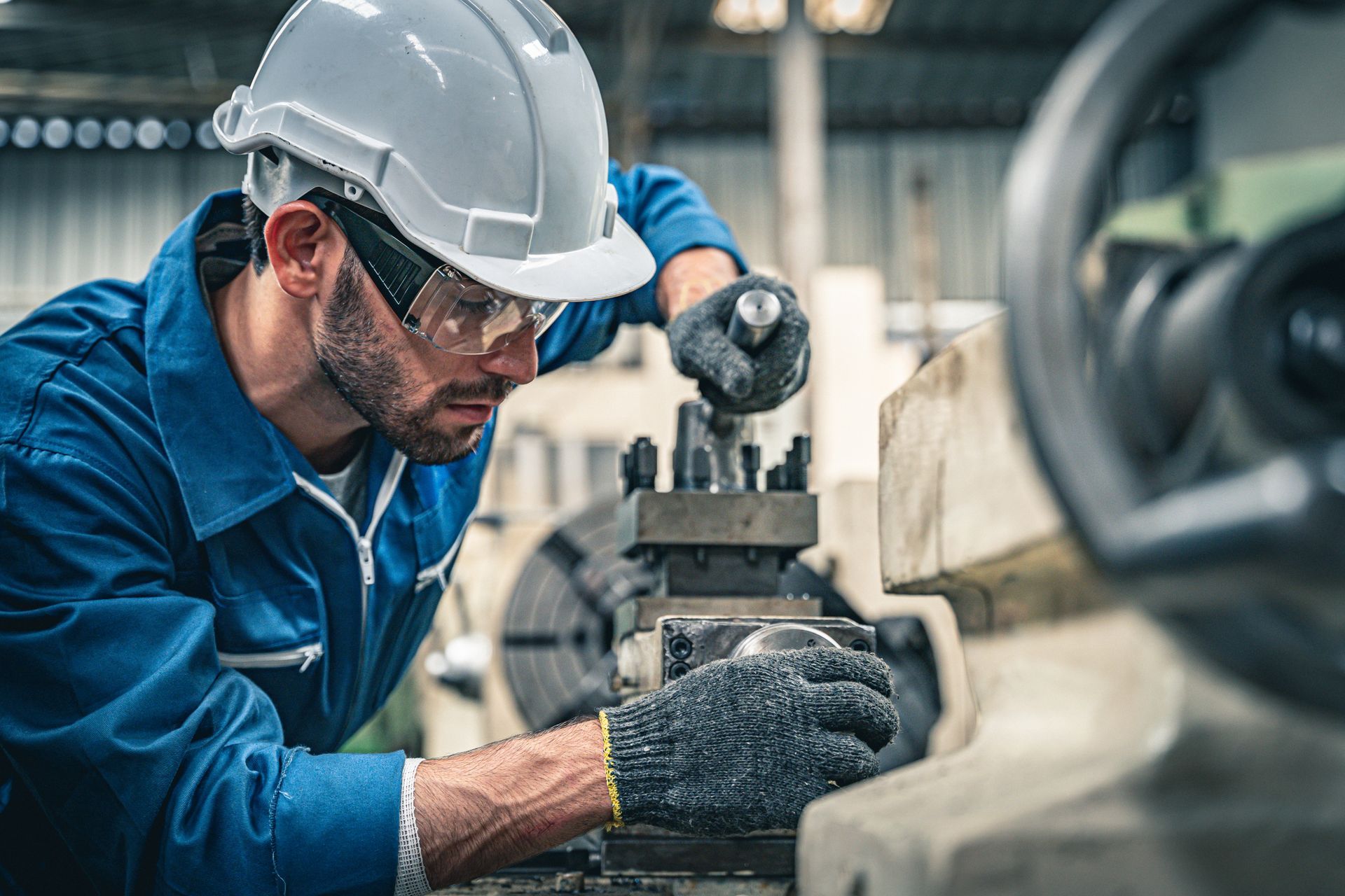 A focused worker in a blue uniform, white hard hat, and safety glasses operates a metal lathe in a factory setting.