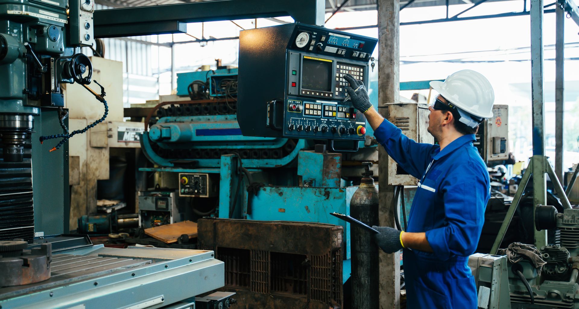 A worker in a blue uniform and hard hat operates a digital control panel on industrial machinery in a factory.