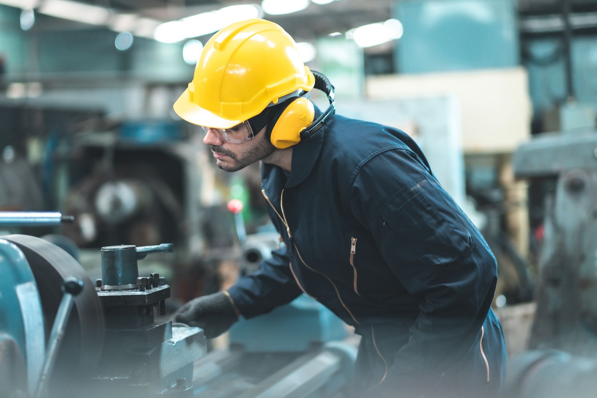 A worker in a yellow hard hat and protective earmuffs operates industrial machinery in a factory.
