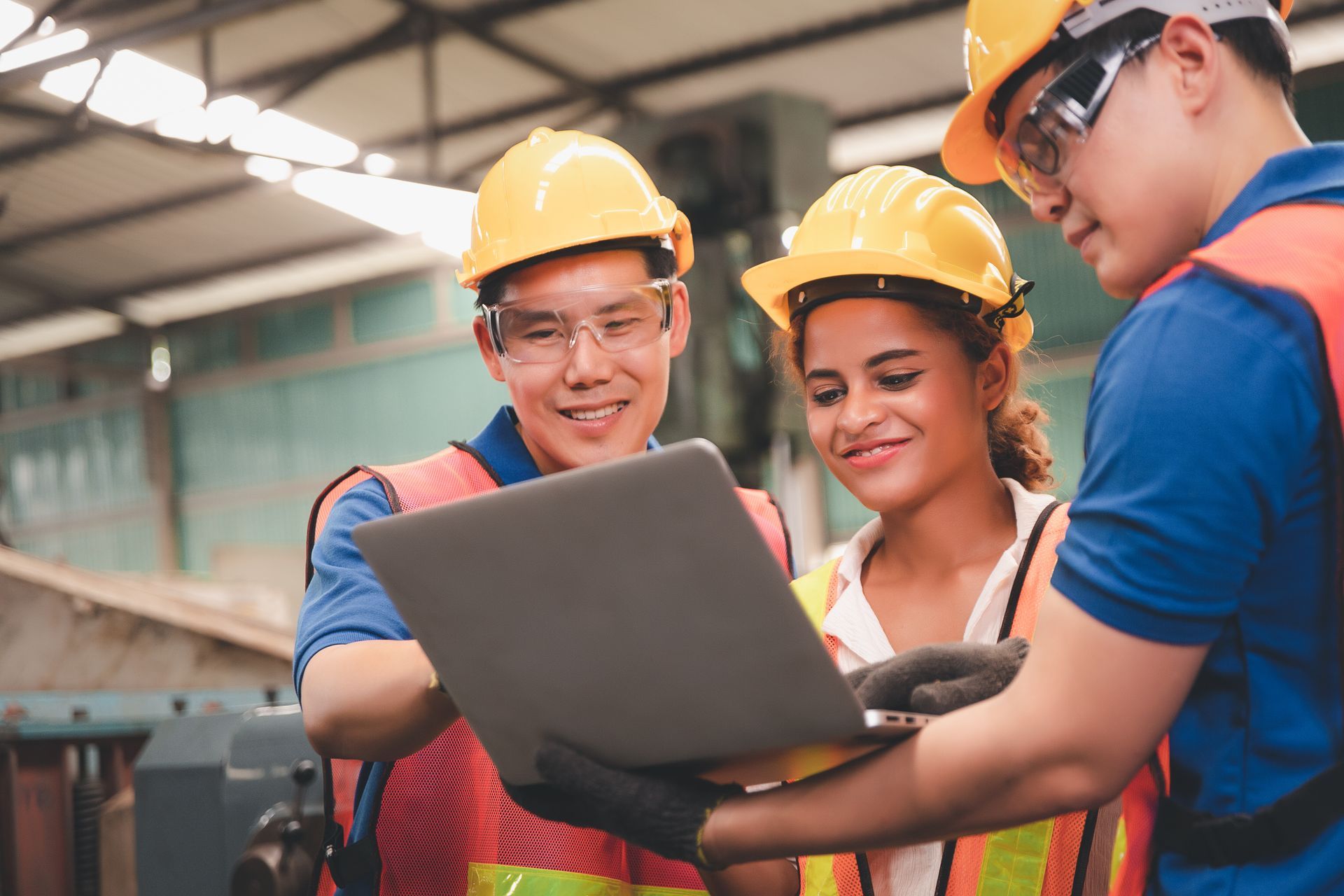 Three construction workers in hard hats and high-visibility vests looking at a laptop in an industrial warehouse setting.