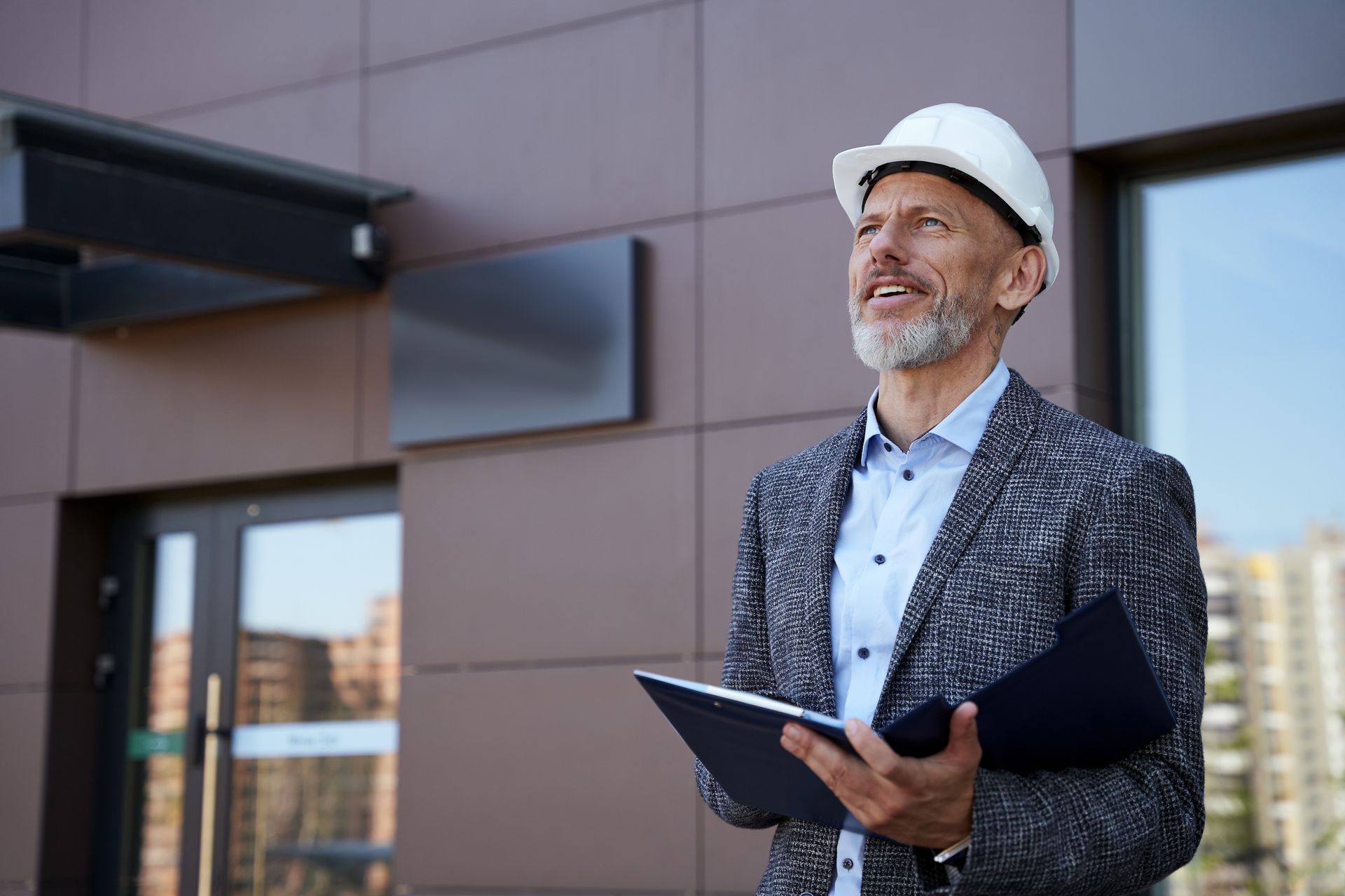 A professional wearing a white hard hat and gray blazer stands outside a building, holding a clipboard and looking up.
