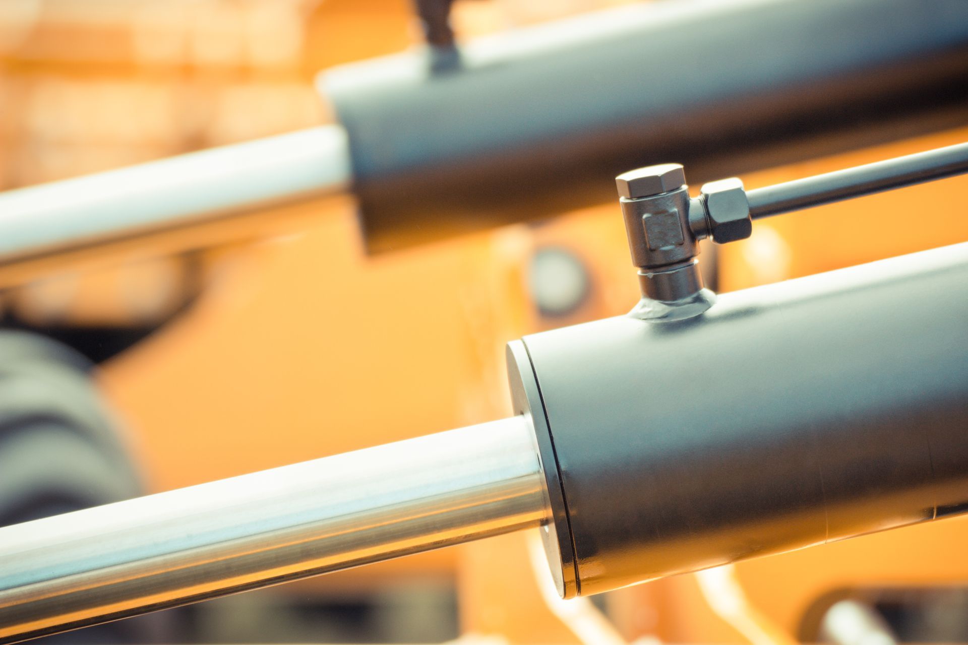 Close-up of matte black hydraulic cylinders with metallic rods and bolted pipe fittings on yellow heavy machinery.