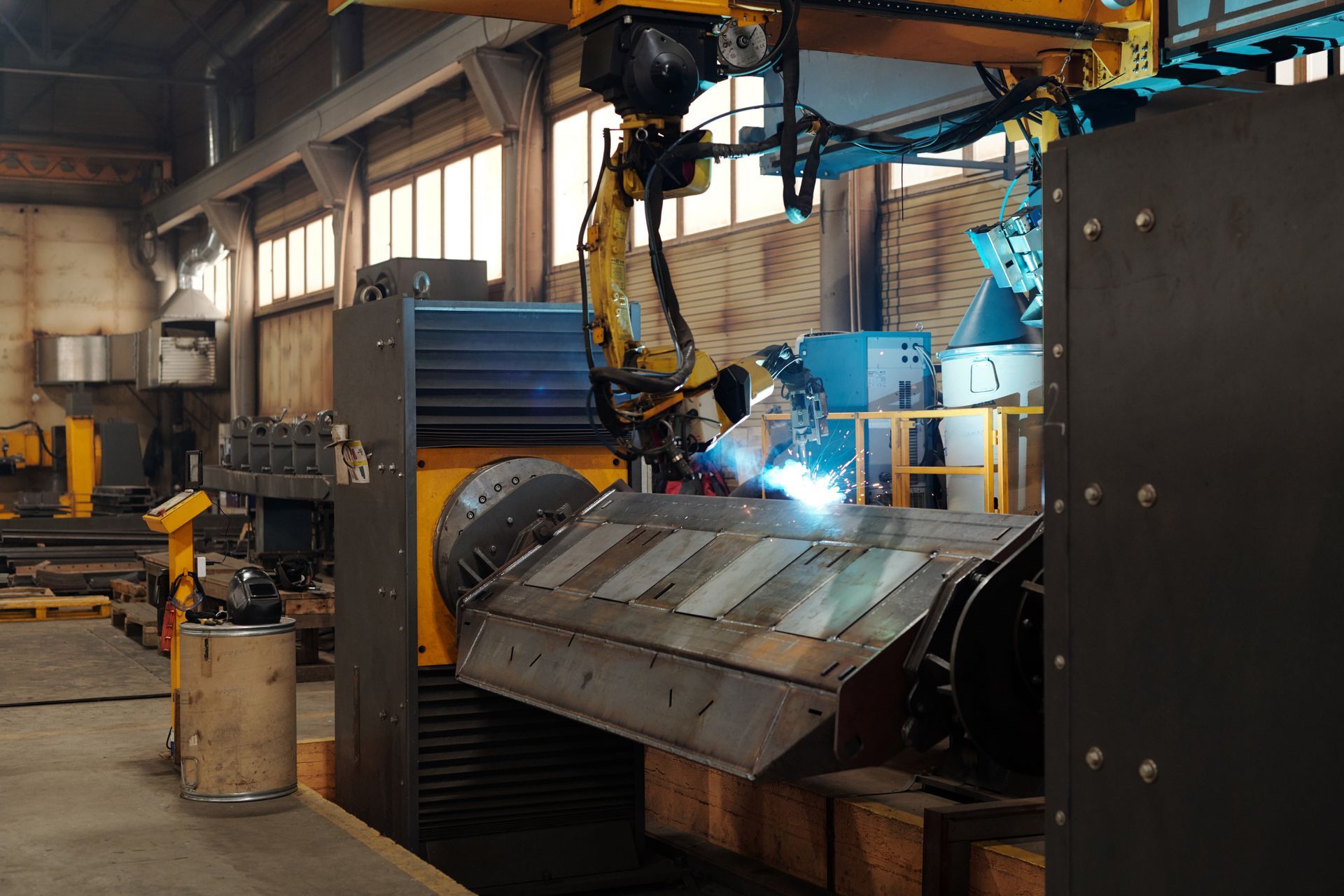 A yellow robotic arm performs arc welding on a large metal workpiece in an industrial factory setting.