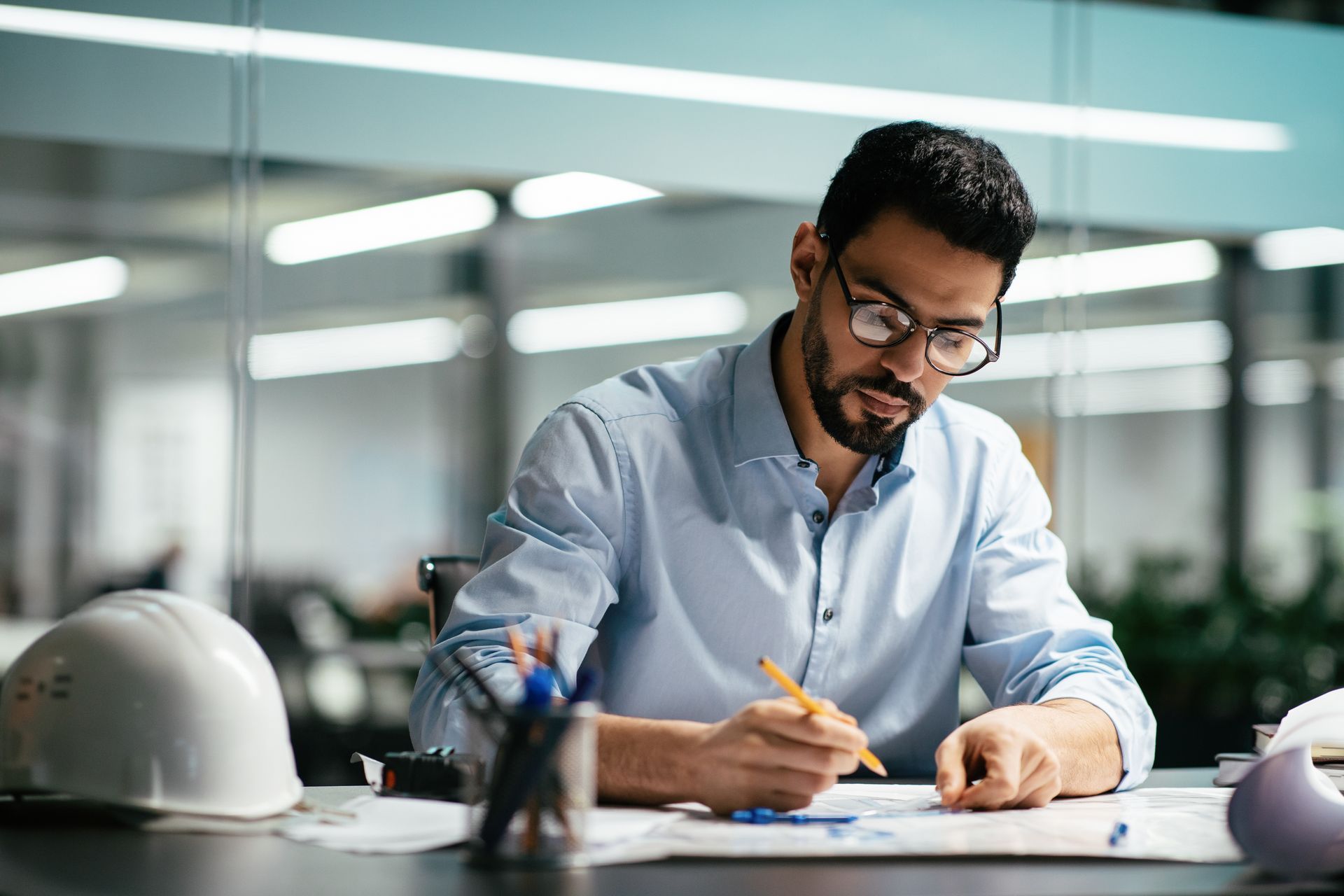 An engineer in a light blue shirt works at a desk, sketching on blueprints next to a white hard hat in an office.