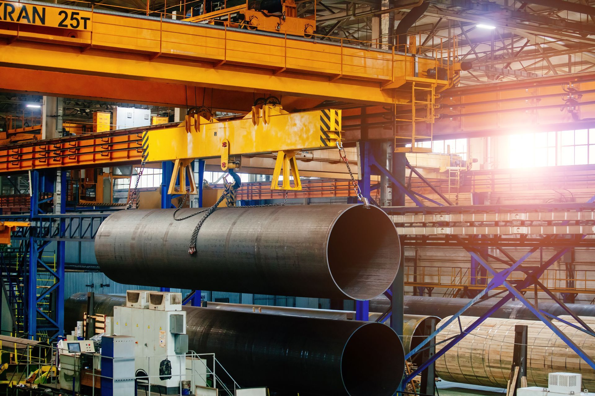 An industrial crane lifts a large, dark steel pipe in a factory setting with bright overhead lighting.