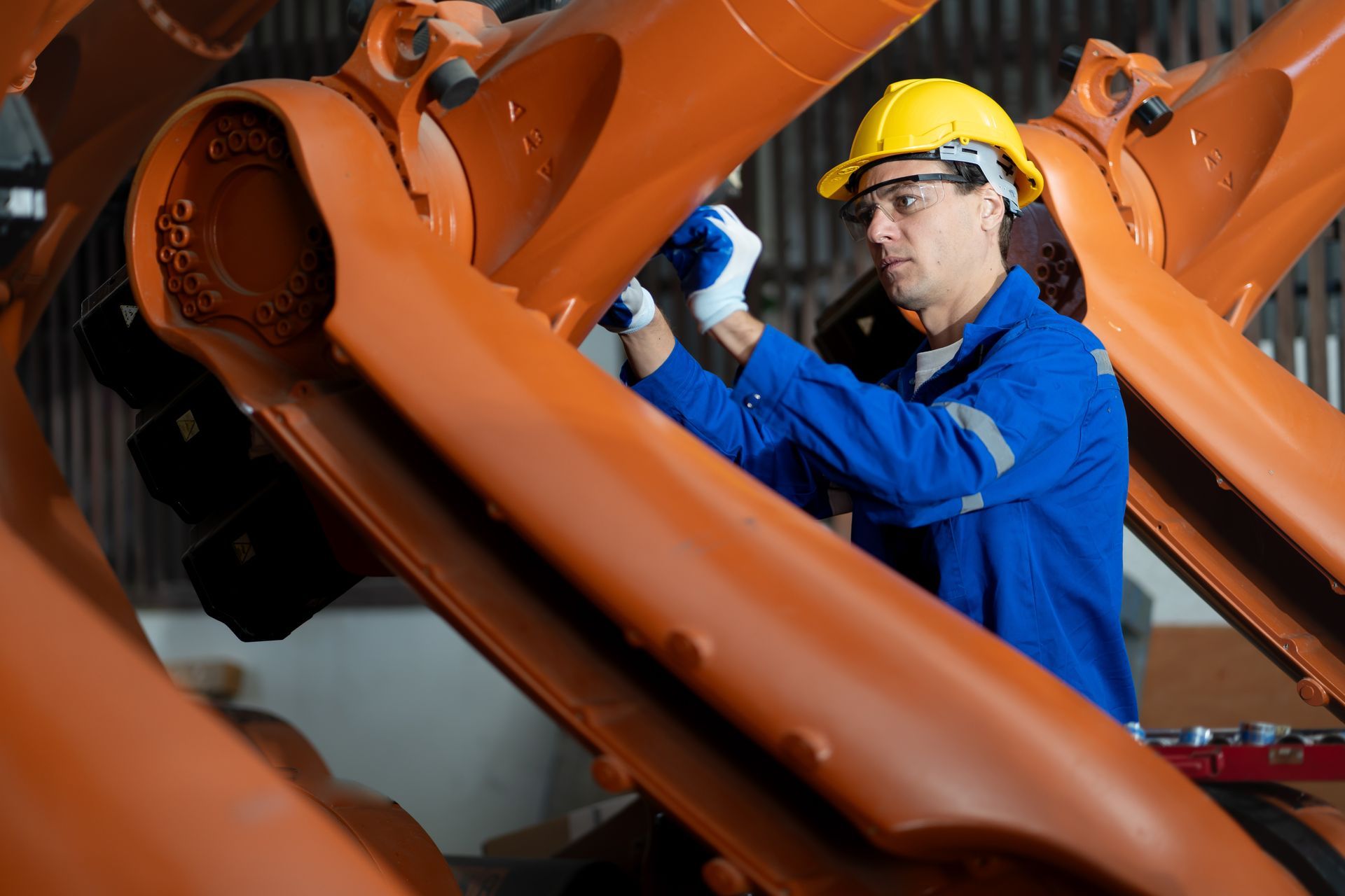 A person in a hard hat and blue uniform works on the mechanical arm of a large orange industrial robot.